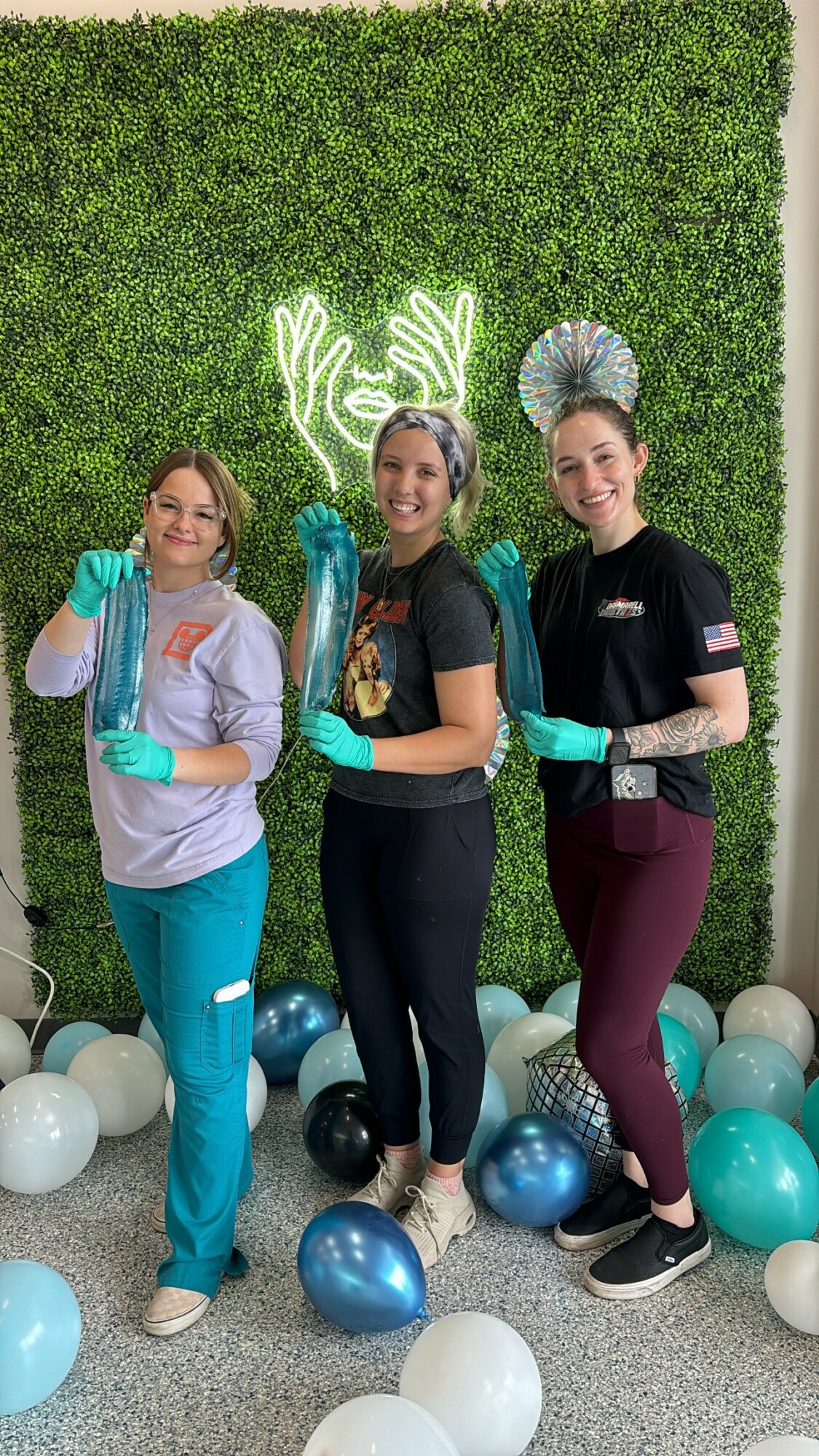 Three women wearing gloves and holding illuminated hand props, standing in front of a green hedge wall with balloons on the floor.