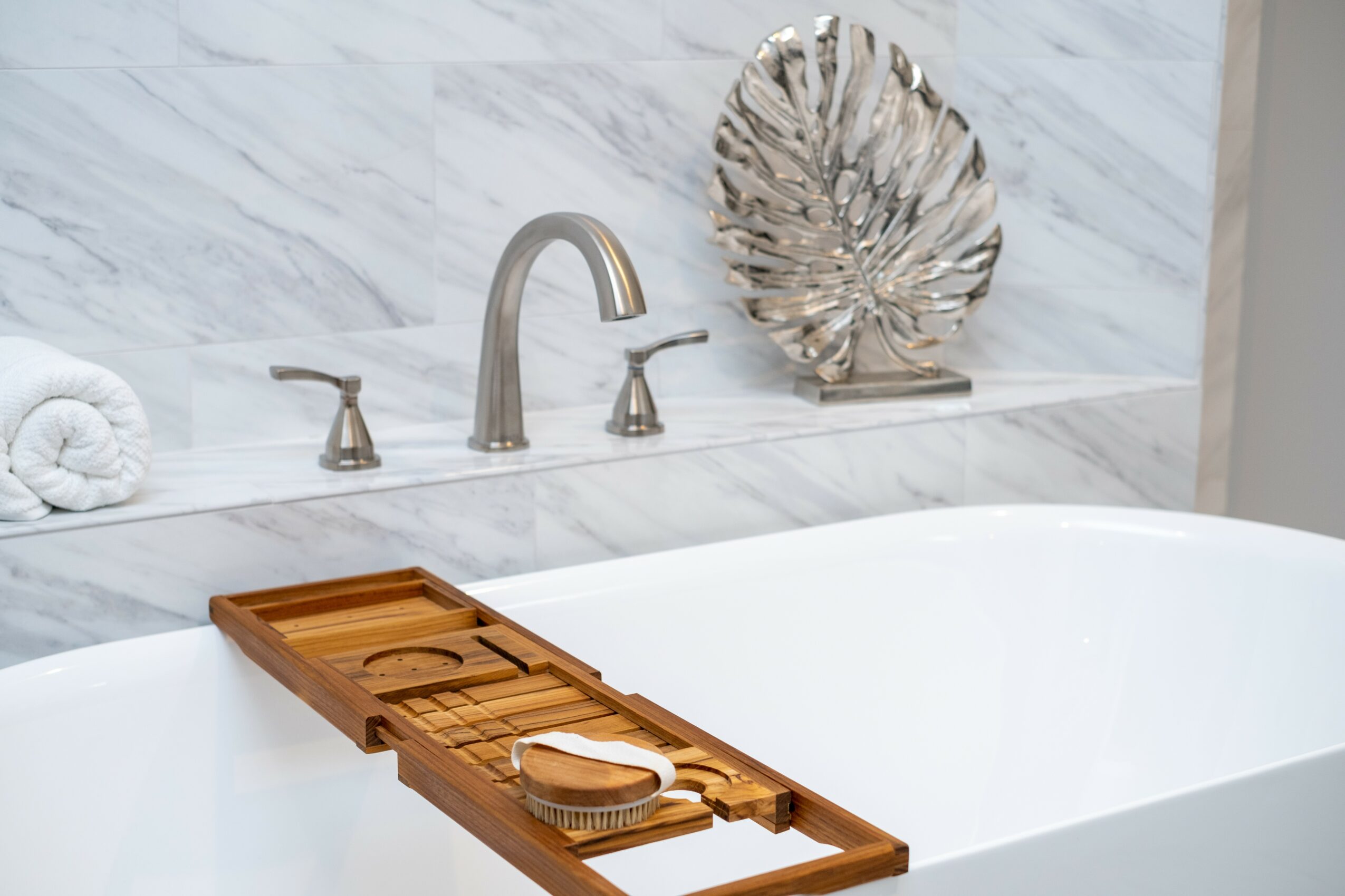 Bathroom sink with a wooden tray holding a soap dish and a small container, marble wall with a decorative shell, faucet, and towel.