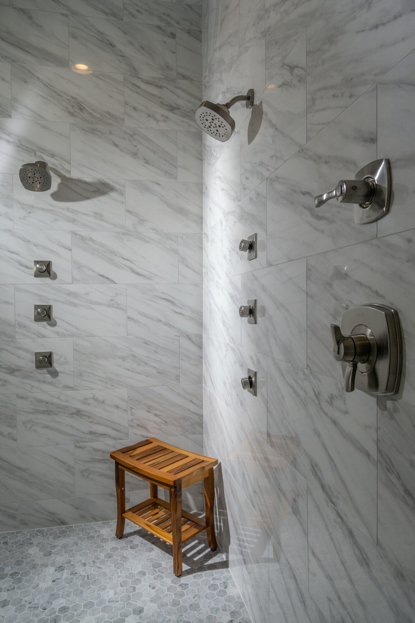 Marble shower with showerhead, controls, a wooden stool, and a marble wall.