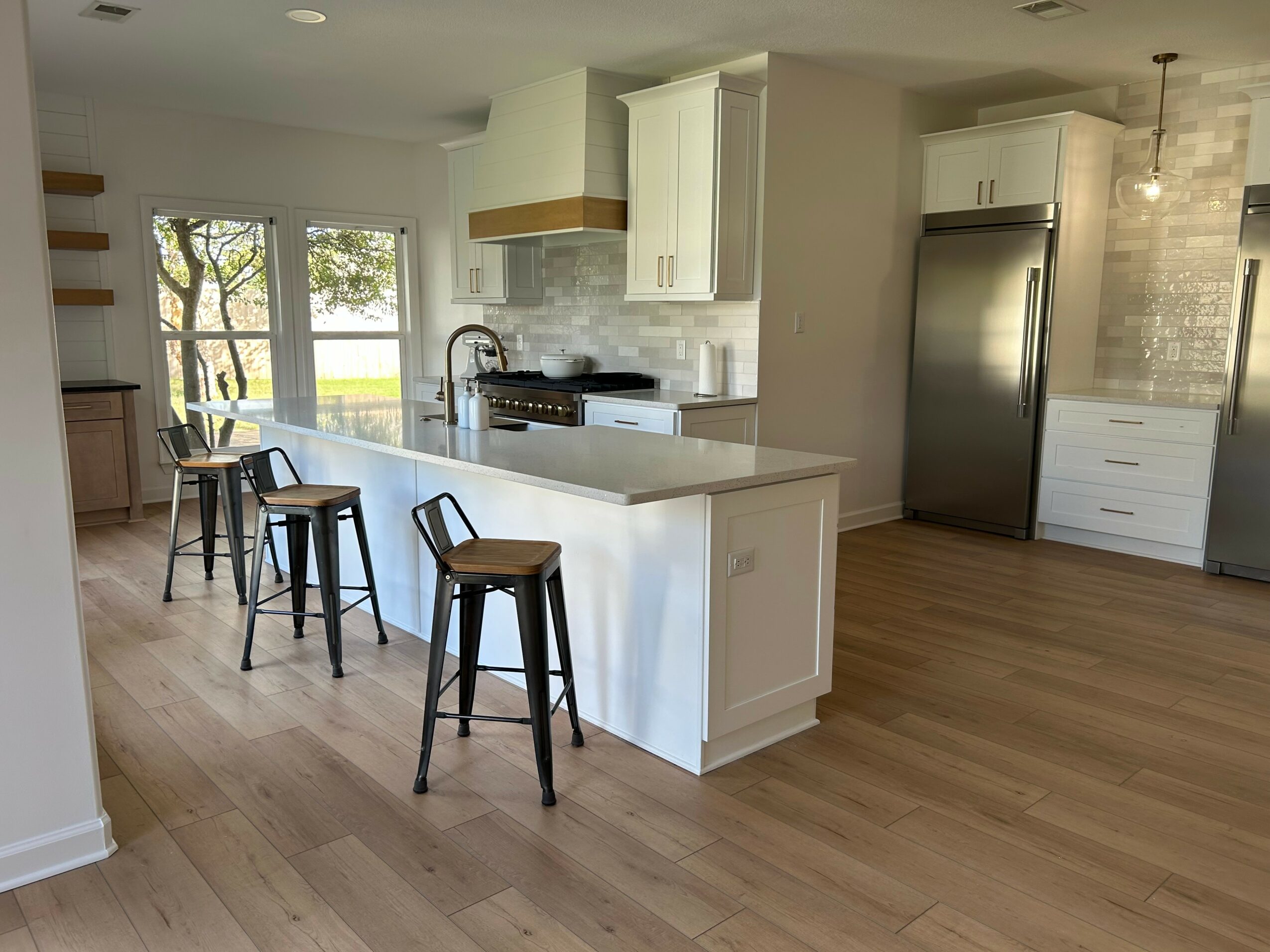Modern kitchen with white cabinets, stainless steel refrigerator, island with three stools, and wooden flooring.