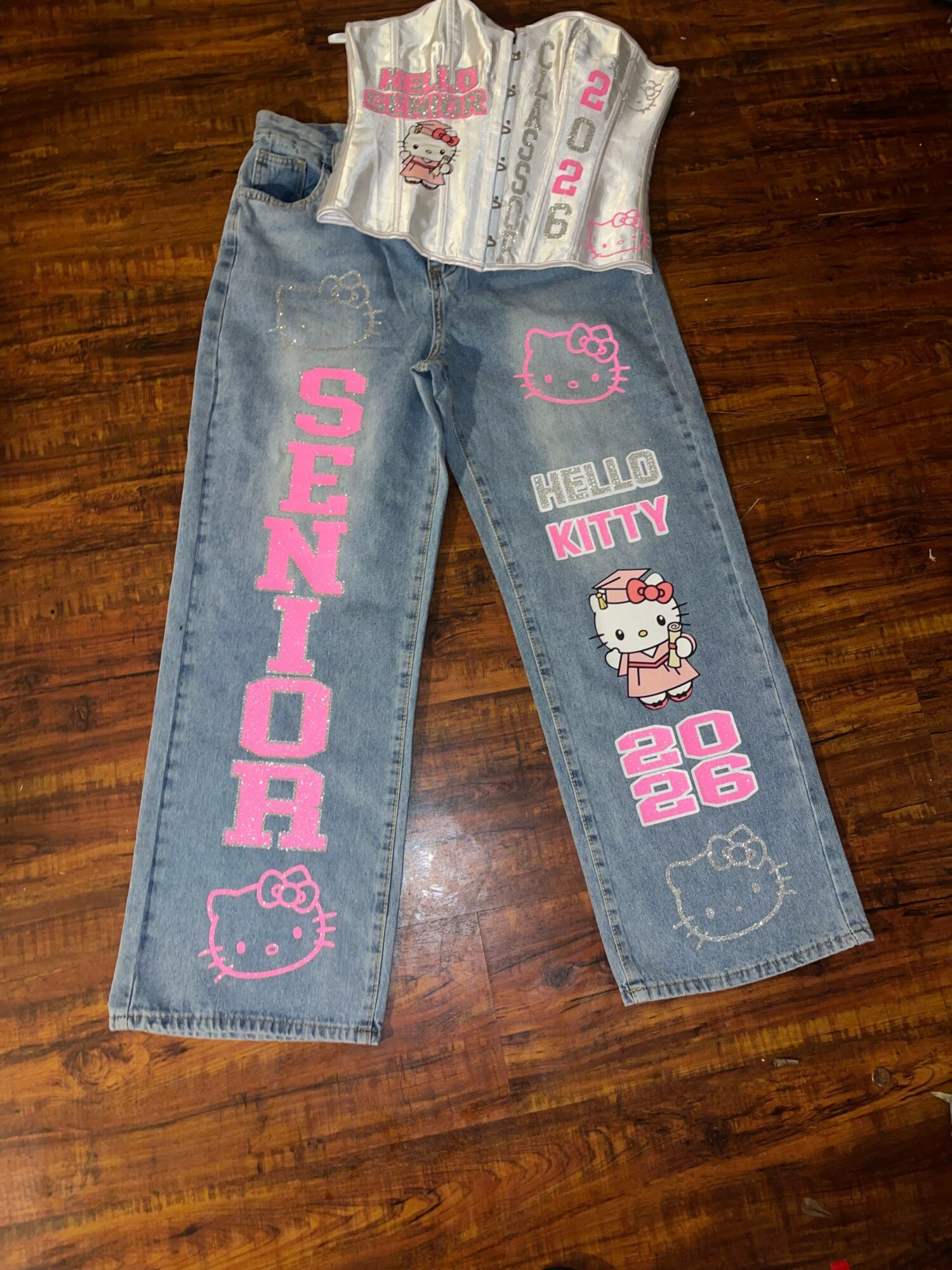 Pair of children's jeans with pink and white Hello Kitty designs and text, laid on a wooden surface.