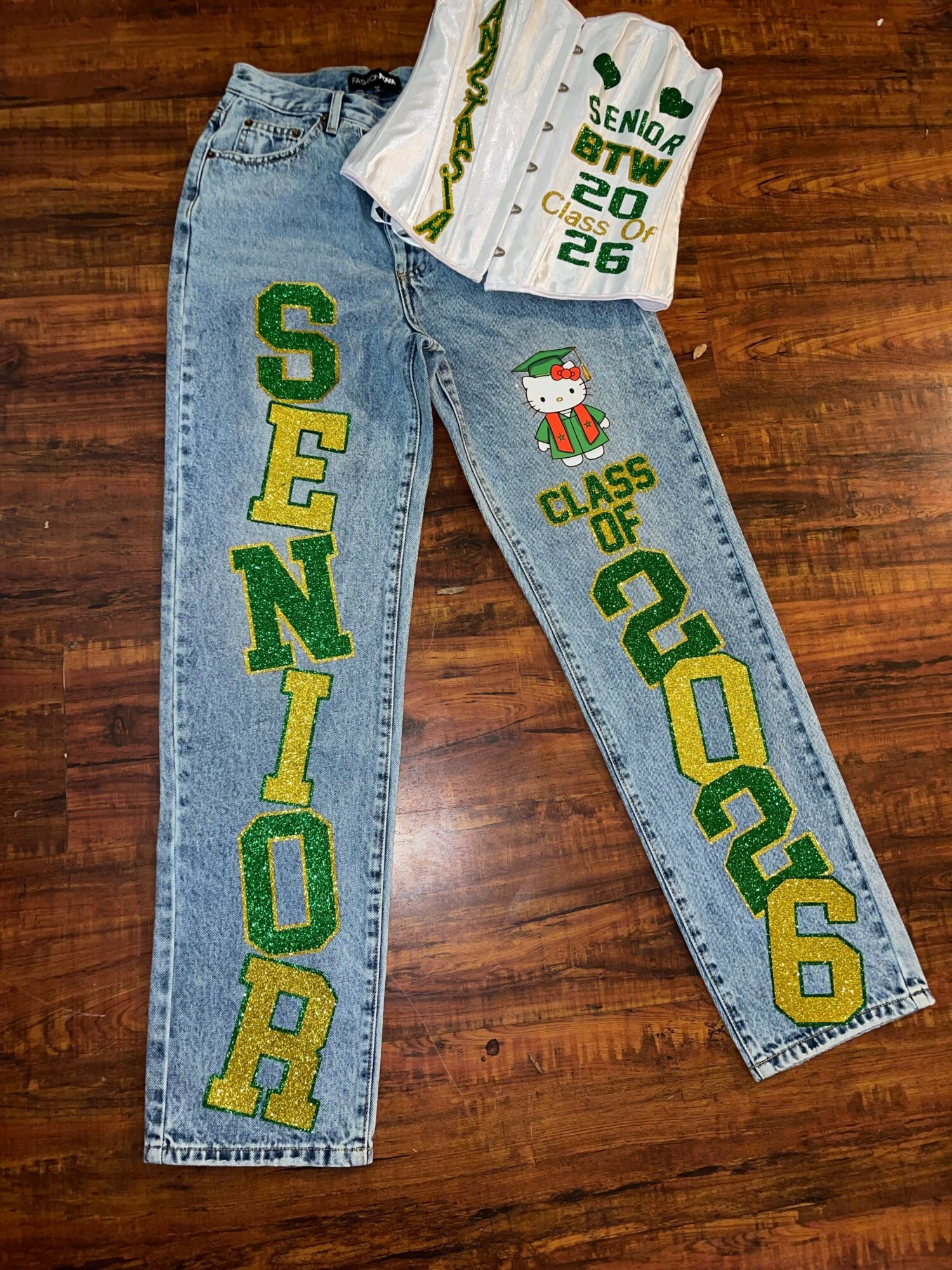 Pair of jeans with green and yellow text and a snowman graphic, laid on a wooden surface, with a white shirt on top.