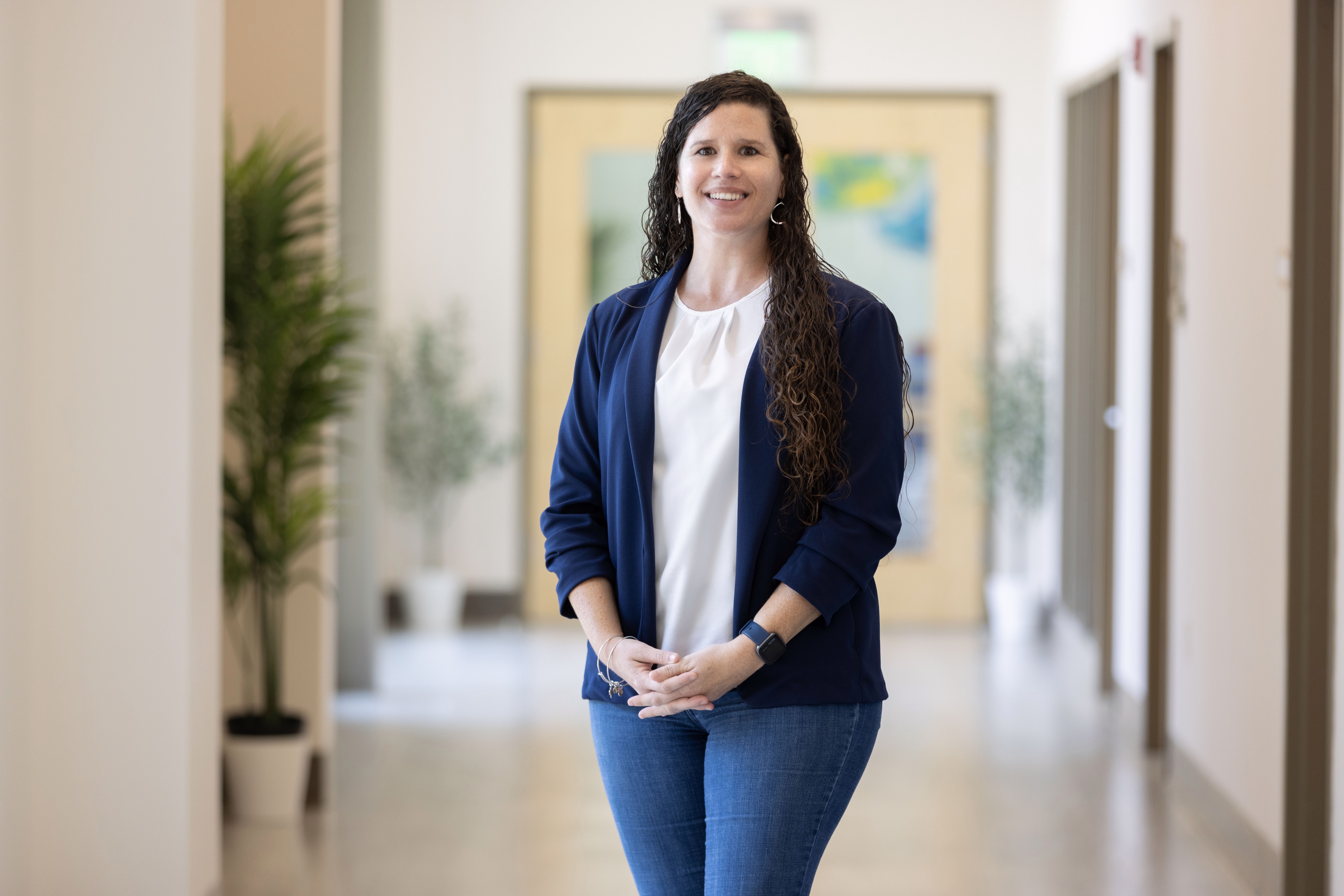Woman with long dark hair smiling, standing in a hallway with plants and artwork in the background.