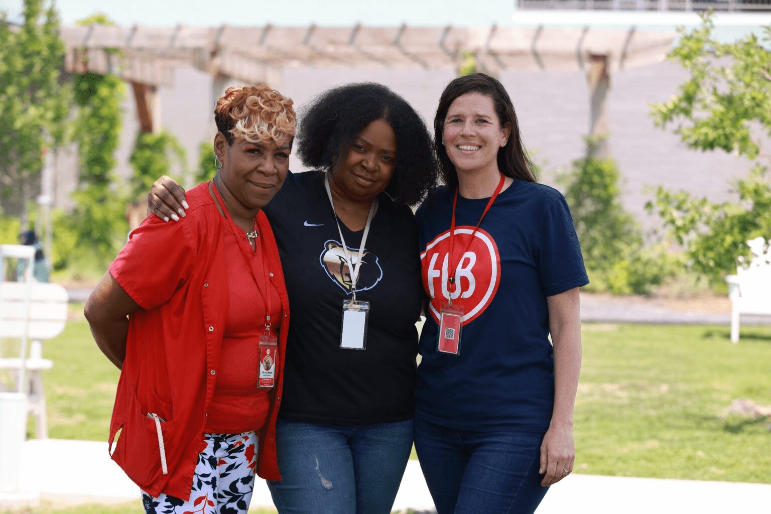 Three women standing outdoors smiling, with trees and a bridge in the background.