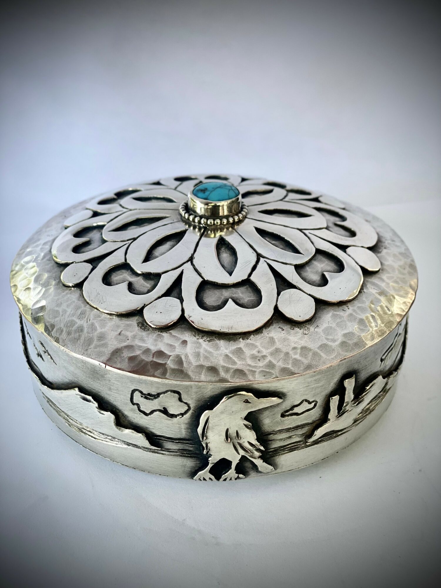 Decorative silver box with intricate floral and animal engravings, topped with a small turquoise and silver ornament.