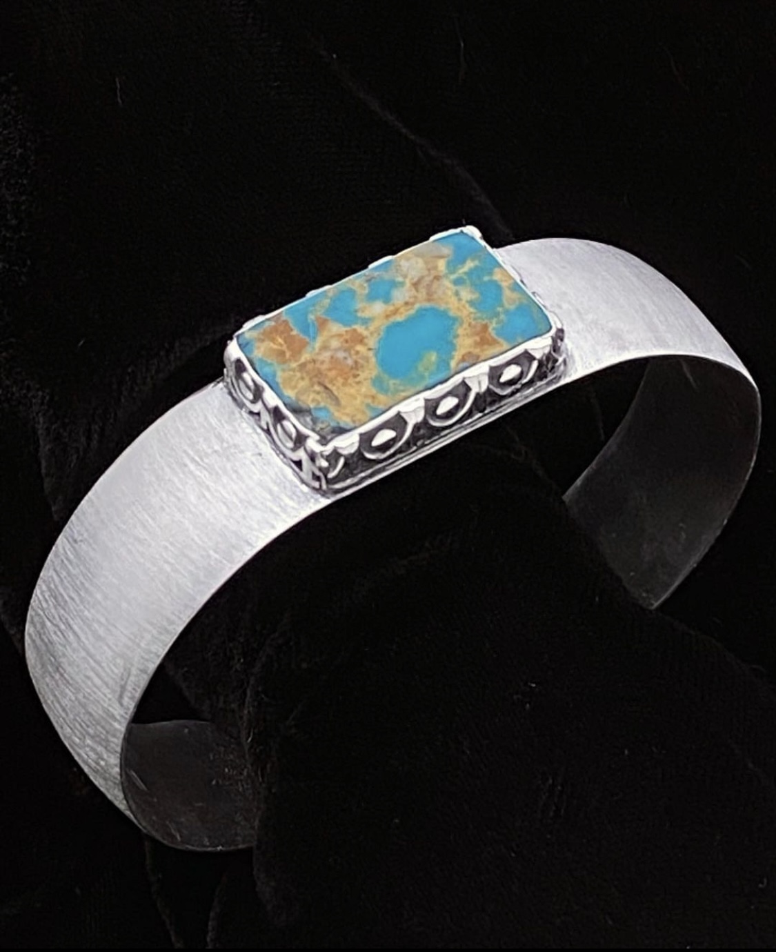 Silver bracelet with a rectangular turquoise stone set in the center, against a black background.