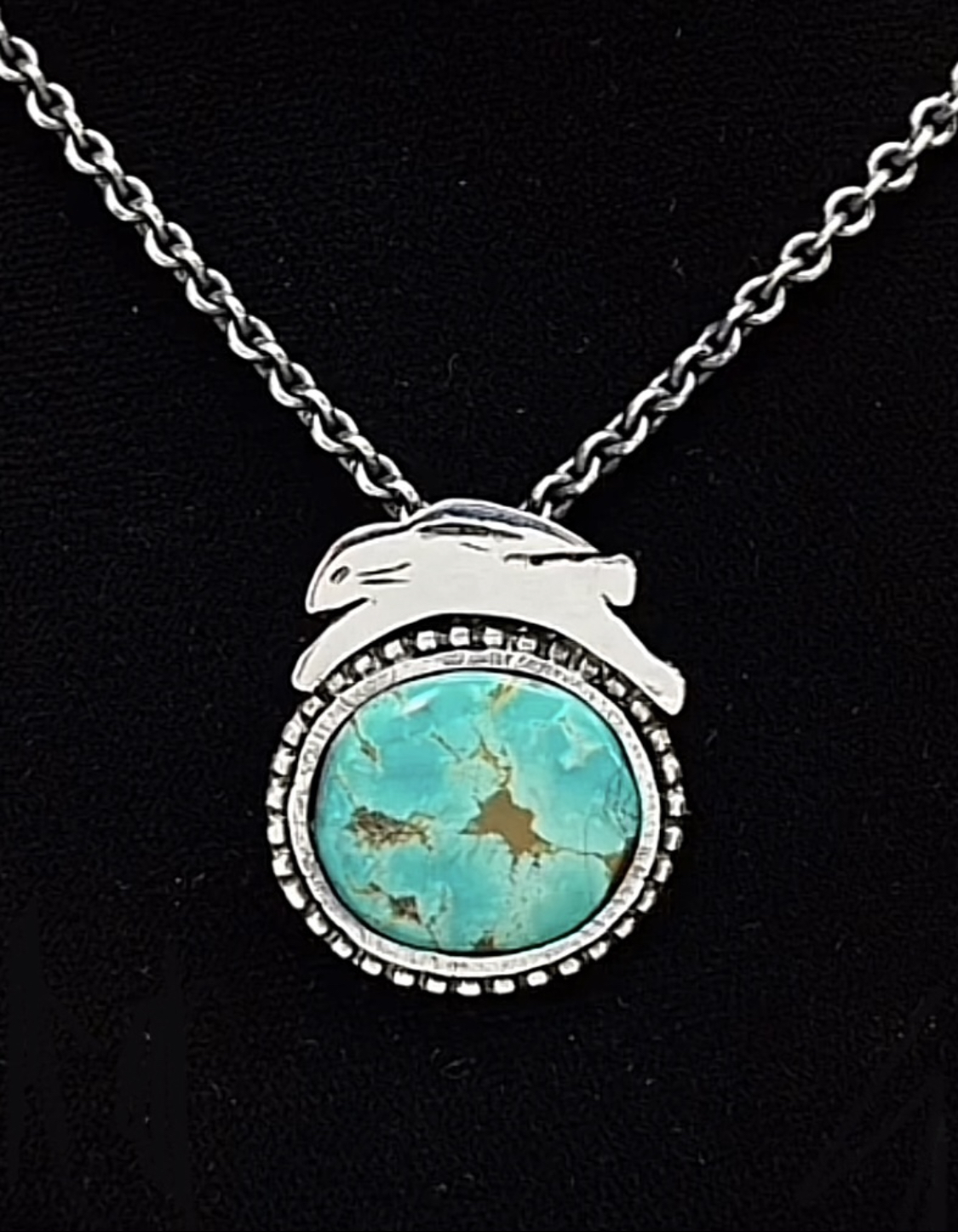 Silver necklace with a round turquoise pendant and a rabbit-shaped detail on top, against a black background.
