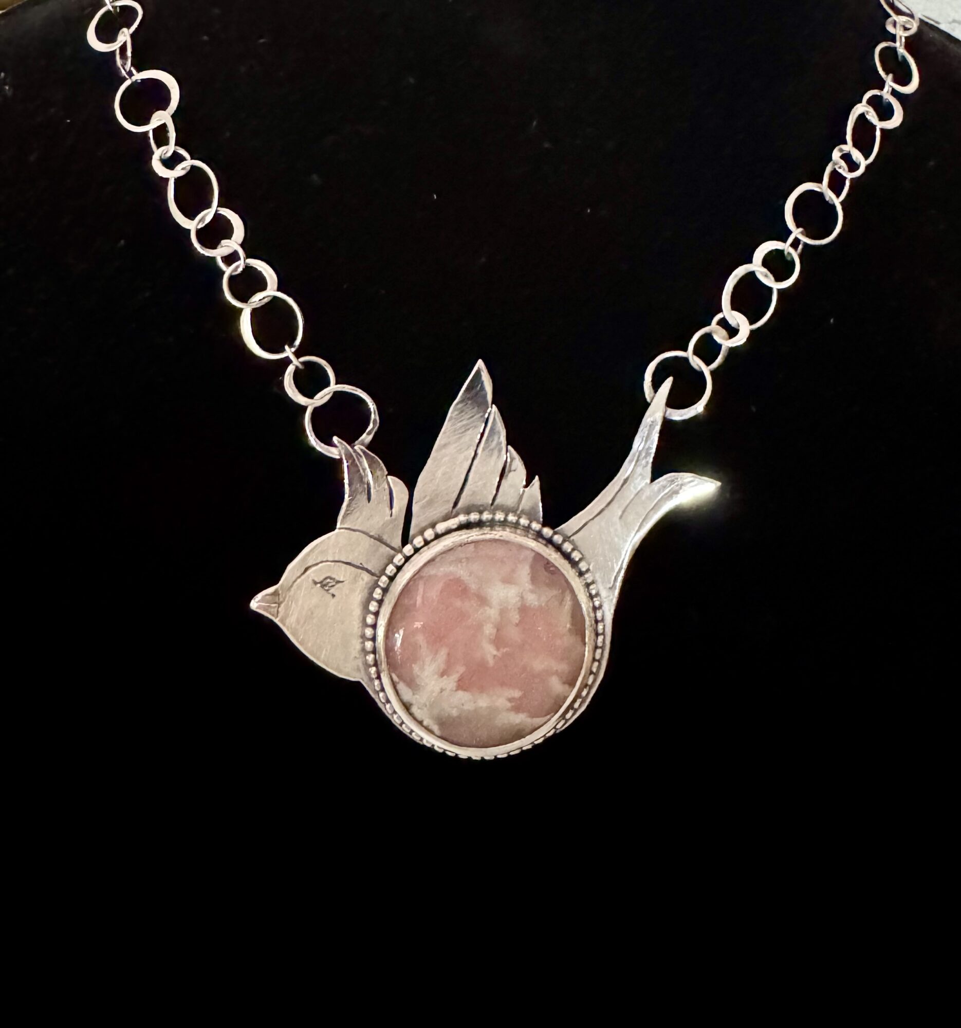 Necklace with a bird-shaped pendant featuring a pink circular stone, on a black background.