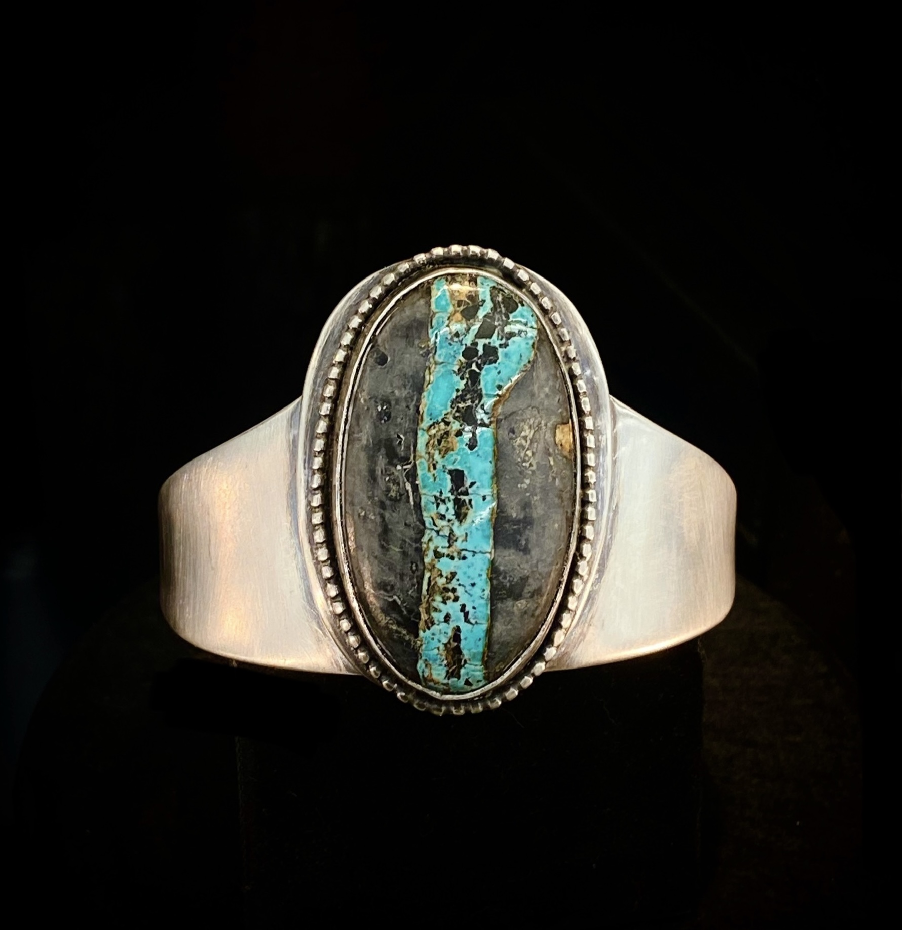 Ring with an oval turquoise and gray stone set in a silver band, detailed border around the stone.