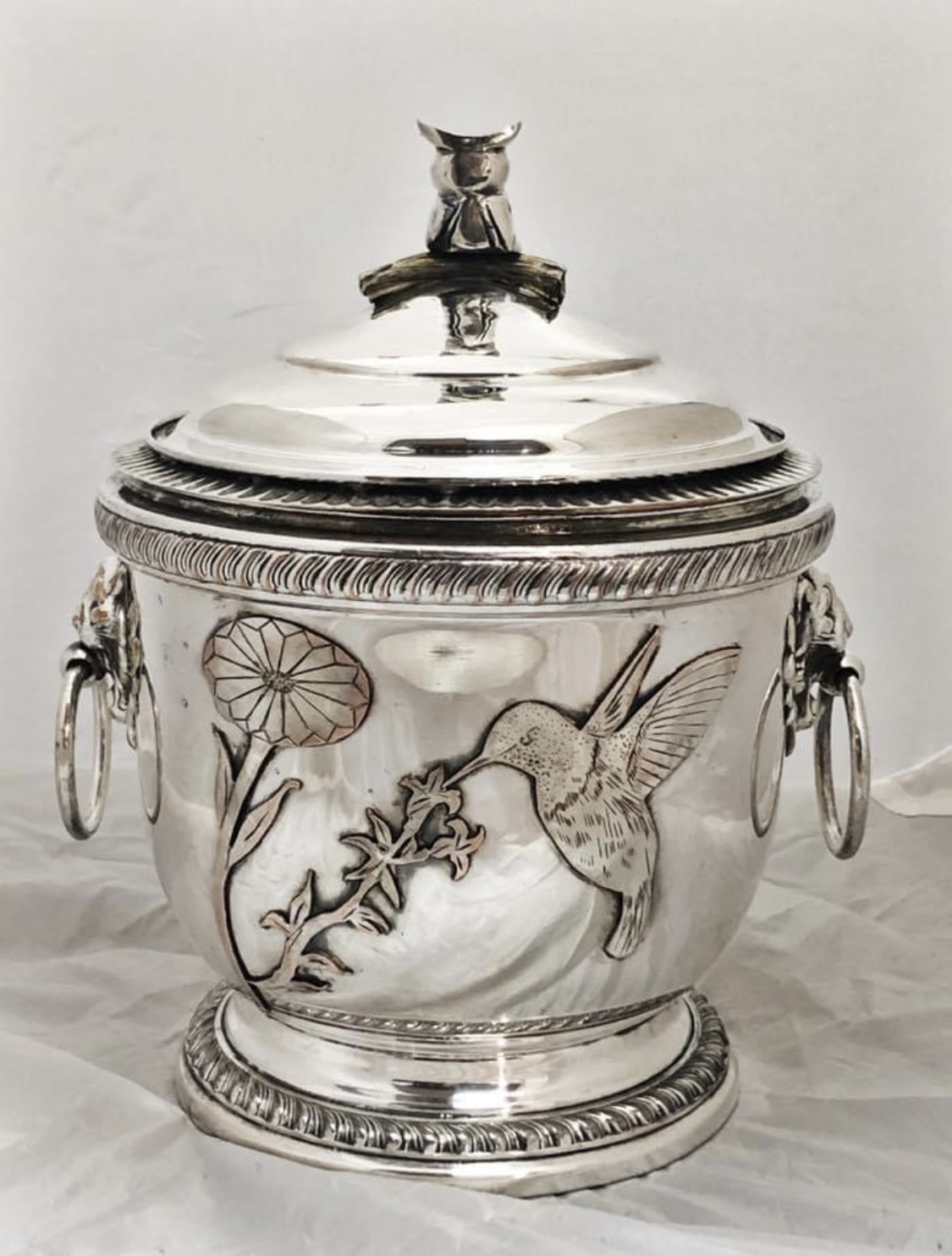 Silver container with a lid, decorated with a hummingbird, flowers, and handles on sides, set against a plain background.