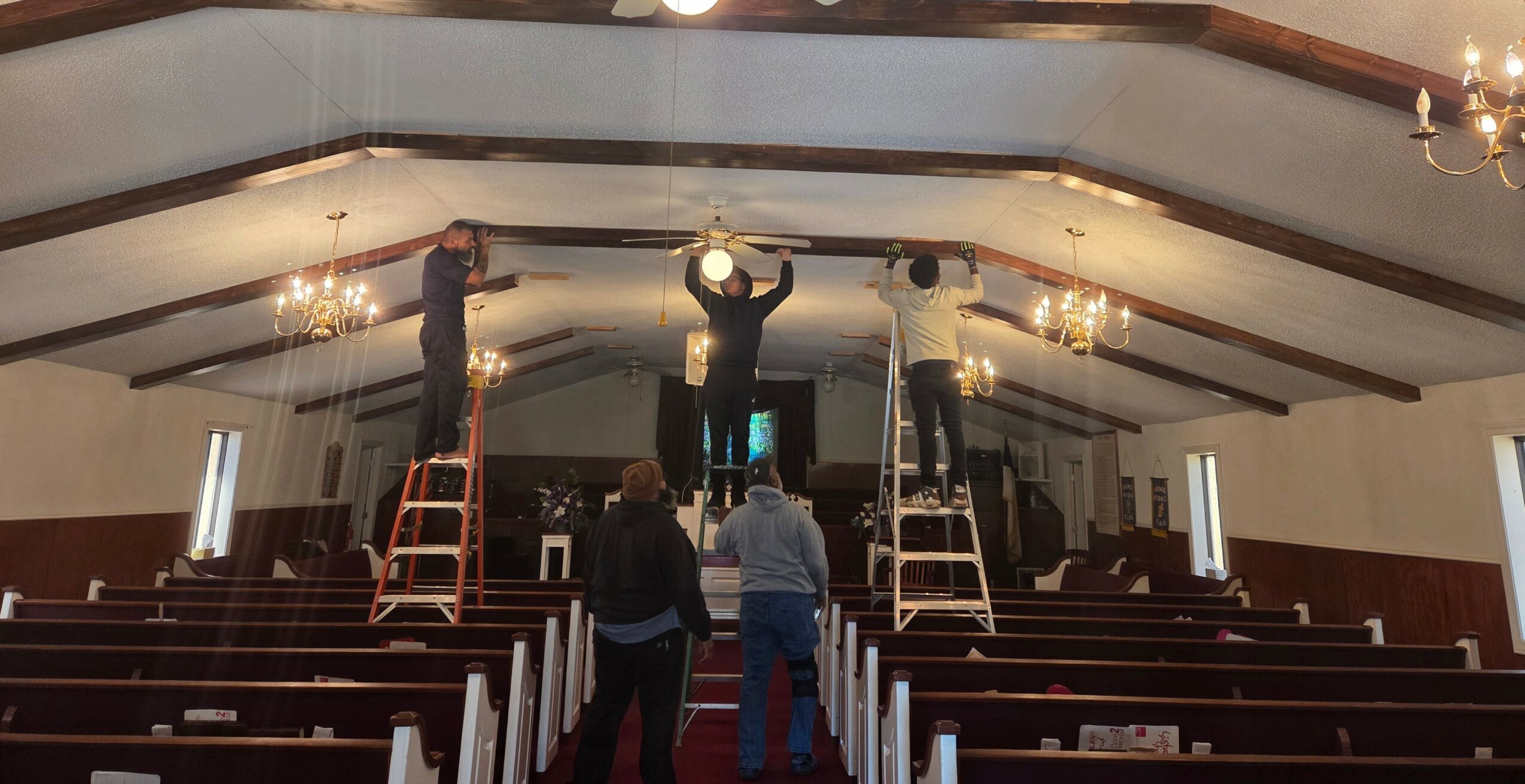 People on ladders working on ceiling lights in a church with wooden pews and chandeliers.