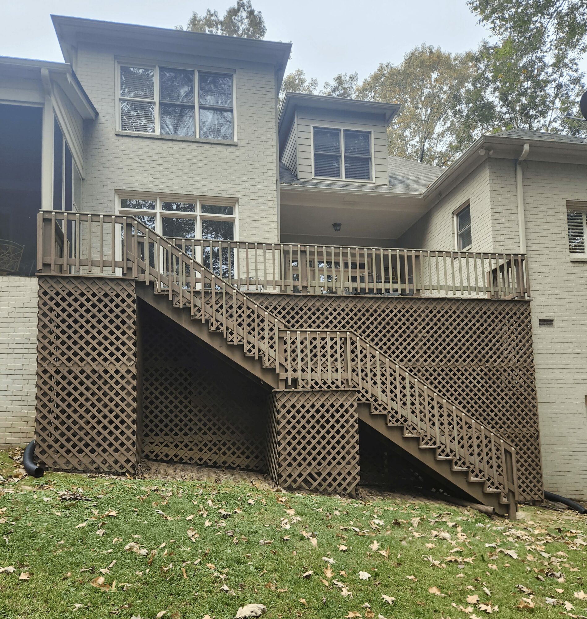 Backyard deck with stairs and lattice, attached to a multi-story house with large windows, trees in background, grass below.
