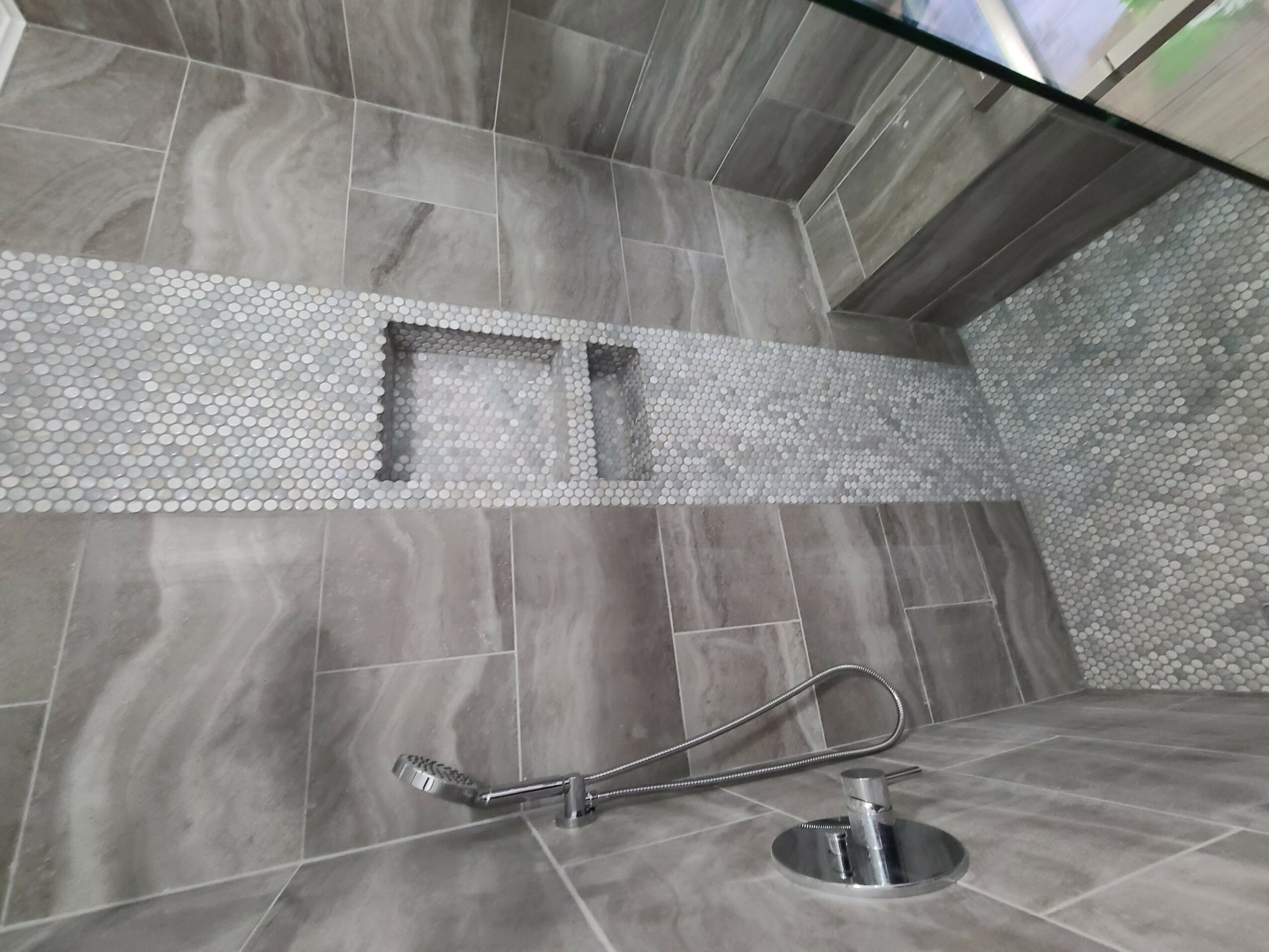 Floor with gray tiles, mosaic tiles, and a metal shower hose on the floor, near a glass wall.