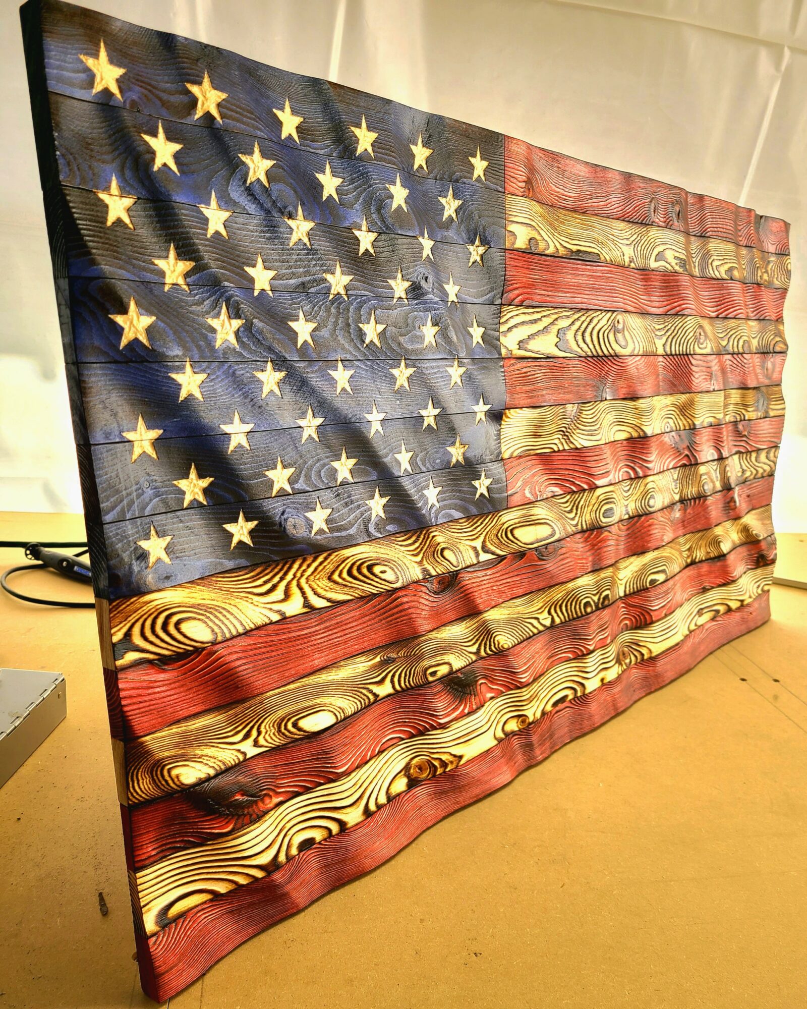 American flag with stars and stripes, hanging vertically, with wood grain pattern on stripes, against a light background.