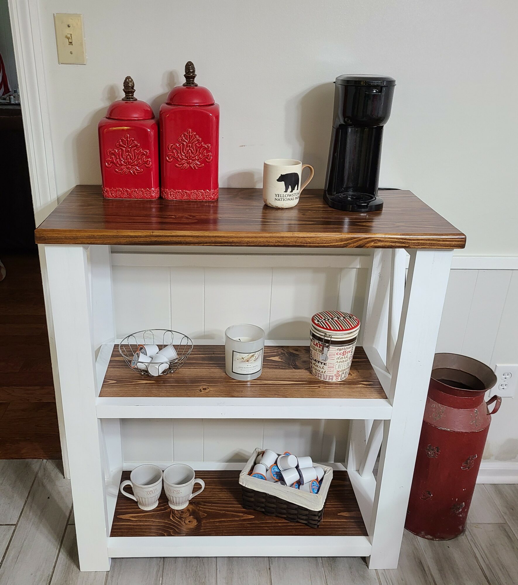 Wooden and white shelf with red containers, a coffee maker, mugs, and a decorative box, against a white wall.