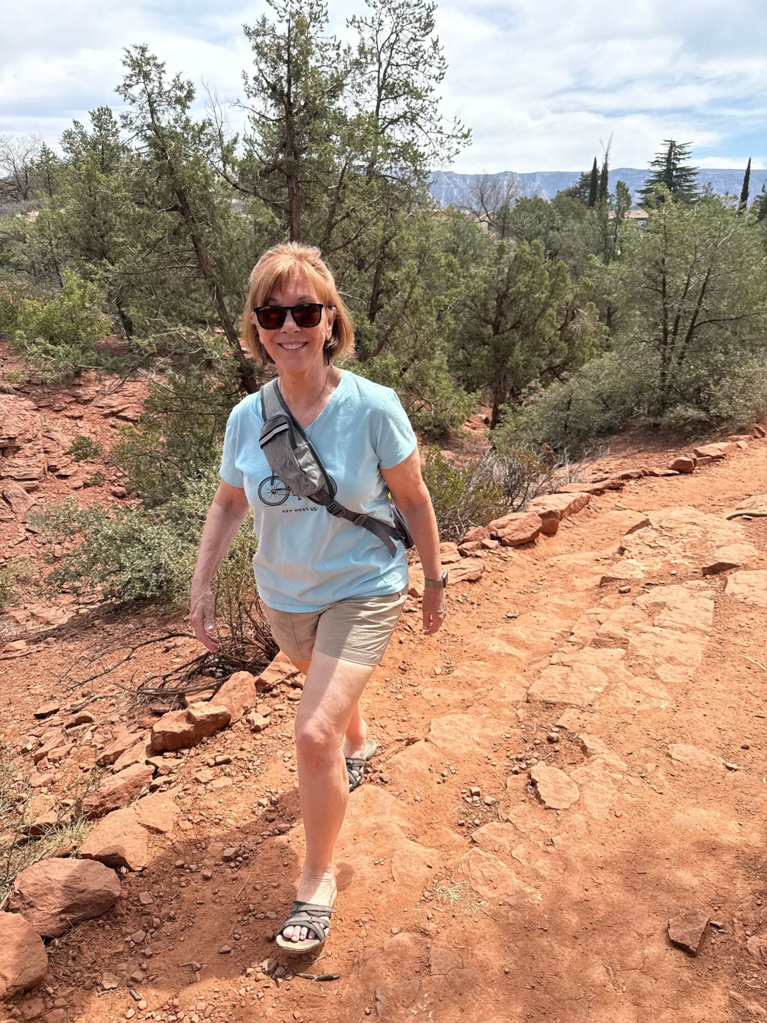 Woman walking on a rocky trail with trees and sky in the background, wearing sunglasses, a light blue shirt, and shorts.