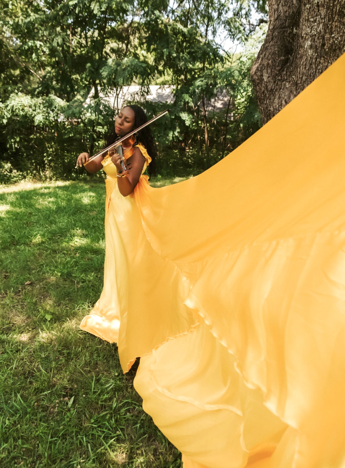Young girl in a flowing yellow dress playing violin outdoors near a tree, with green grass and trees in background.