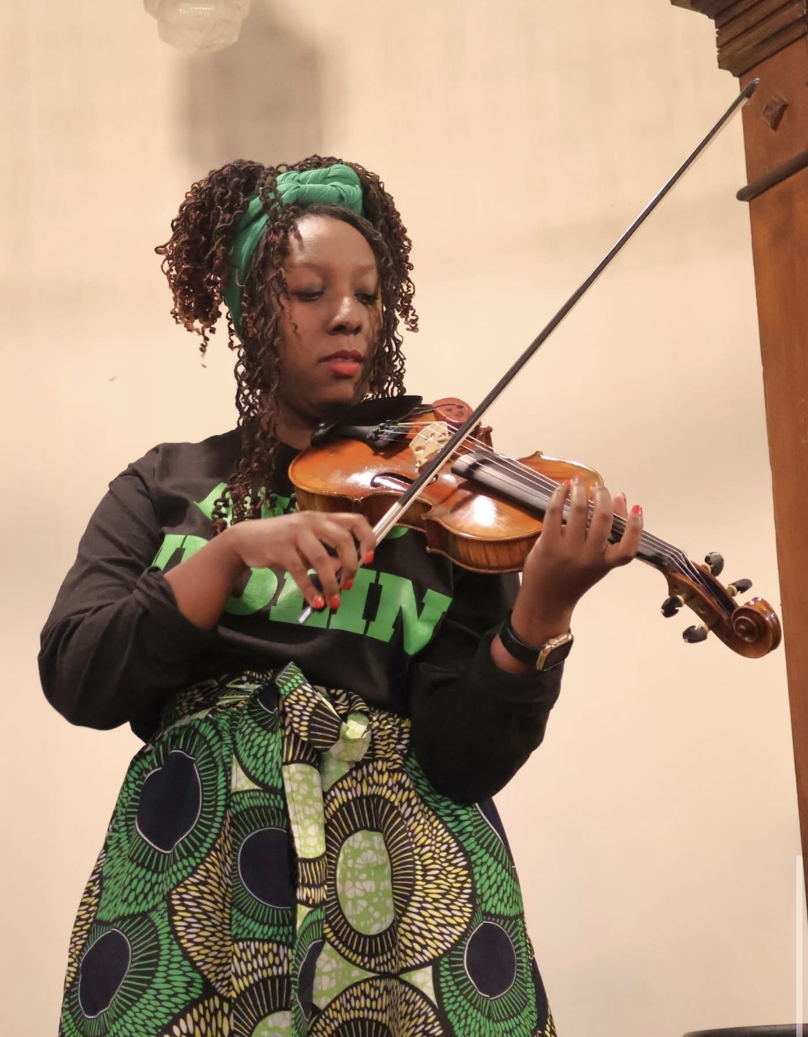 Young girl with curly hair, wearing a green headband, playing a violin, dressed in black and green clothing, standing indoors.