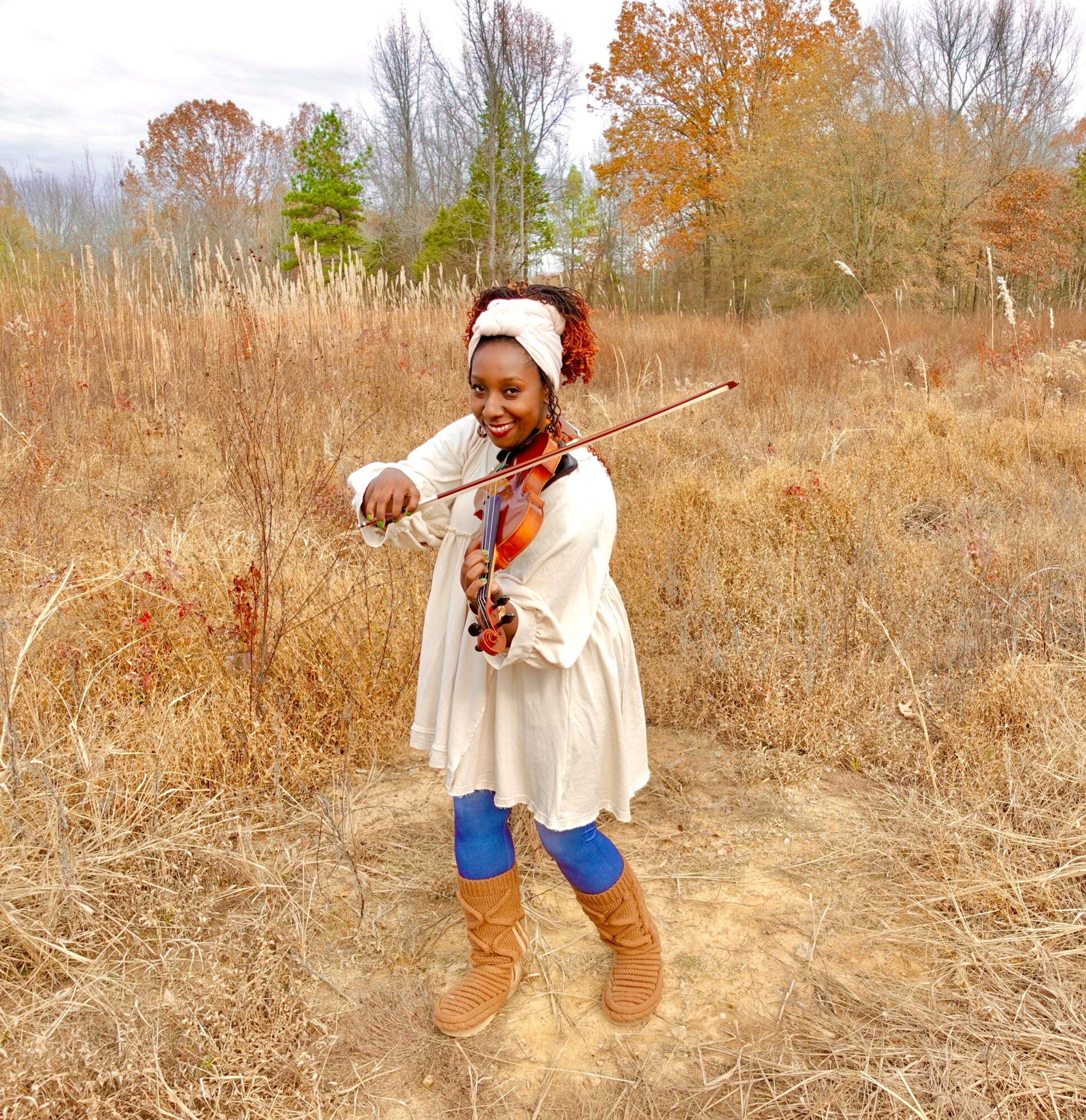 Girl playing violin outdoors in a field with trees and autumn foliage, smiling, wearing a white dress, blue leggings, and brown boots.