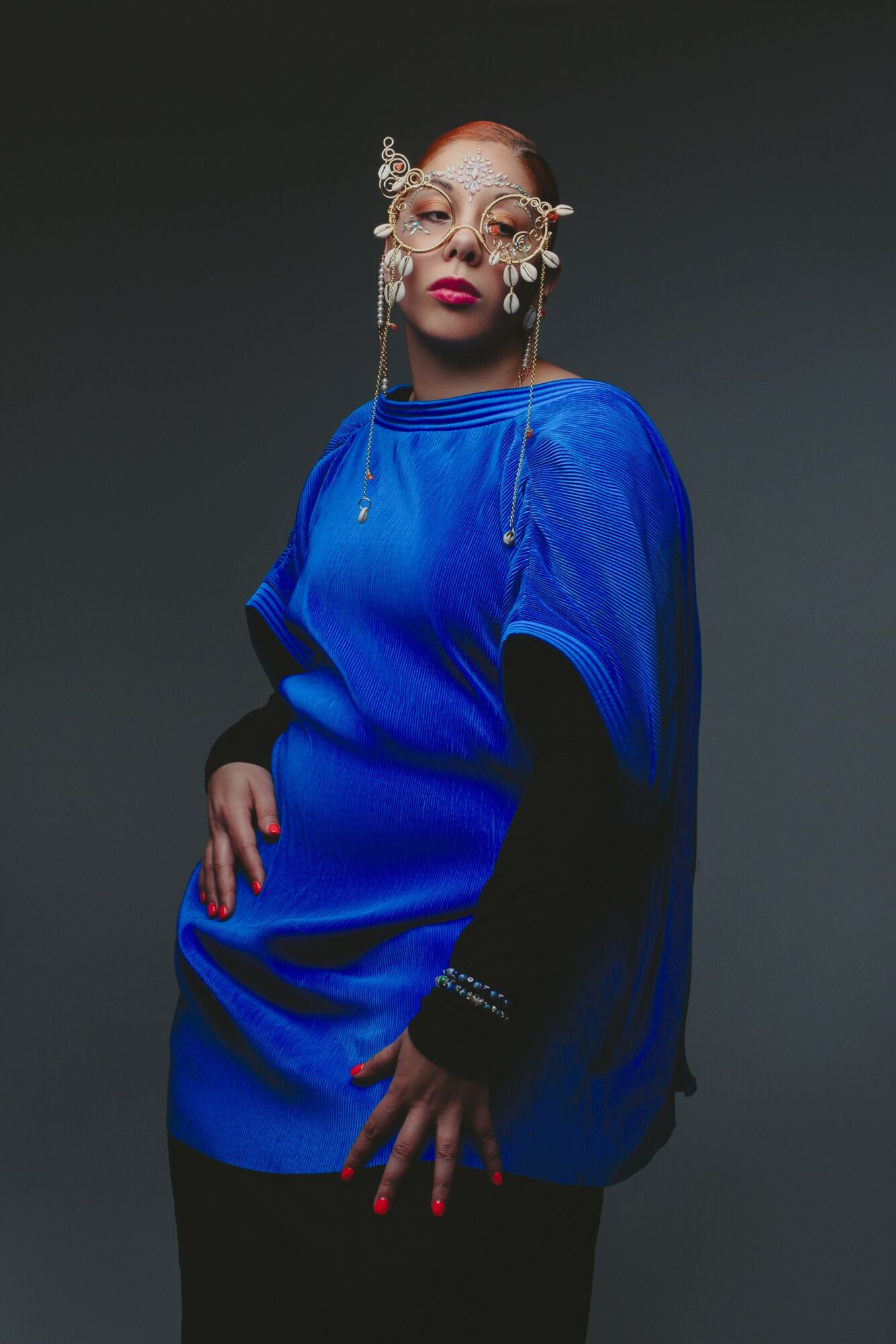 Person with elaborate glasses and bold makeup wearing a blue oversized sweater against a dark background.