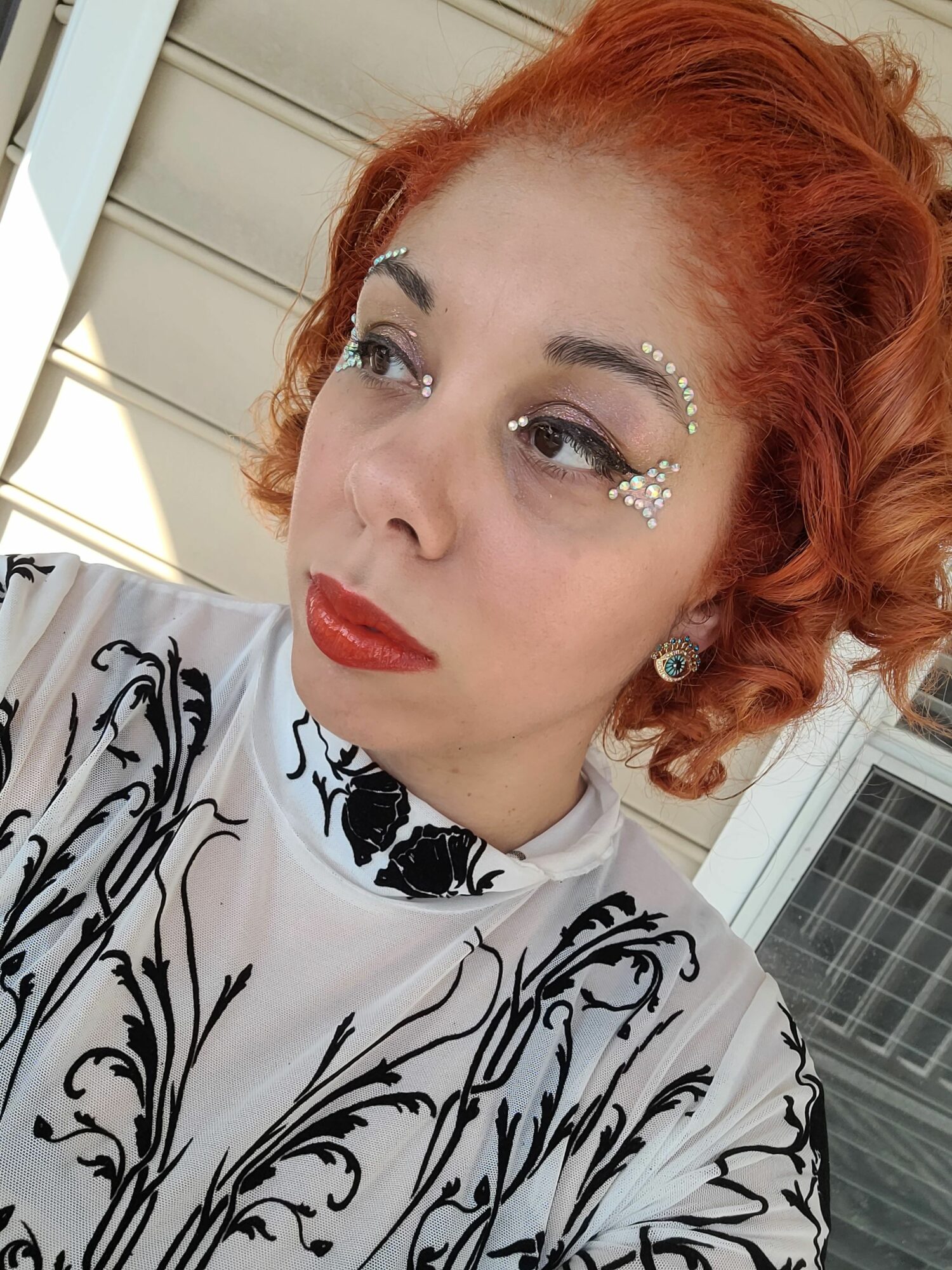 Close-up of a woman with red curly hair, makeup, and decorative face pearls, wearing a patterned top, looking to the side.