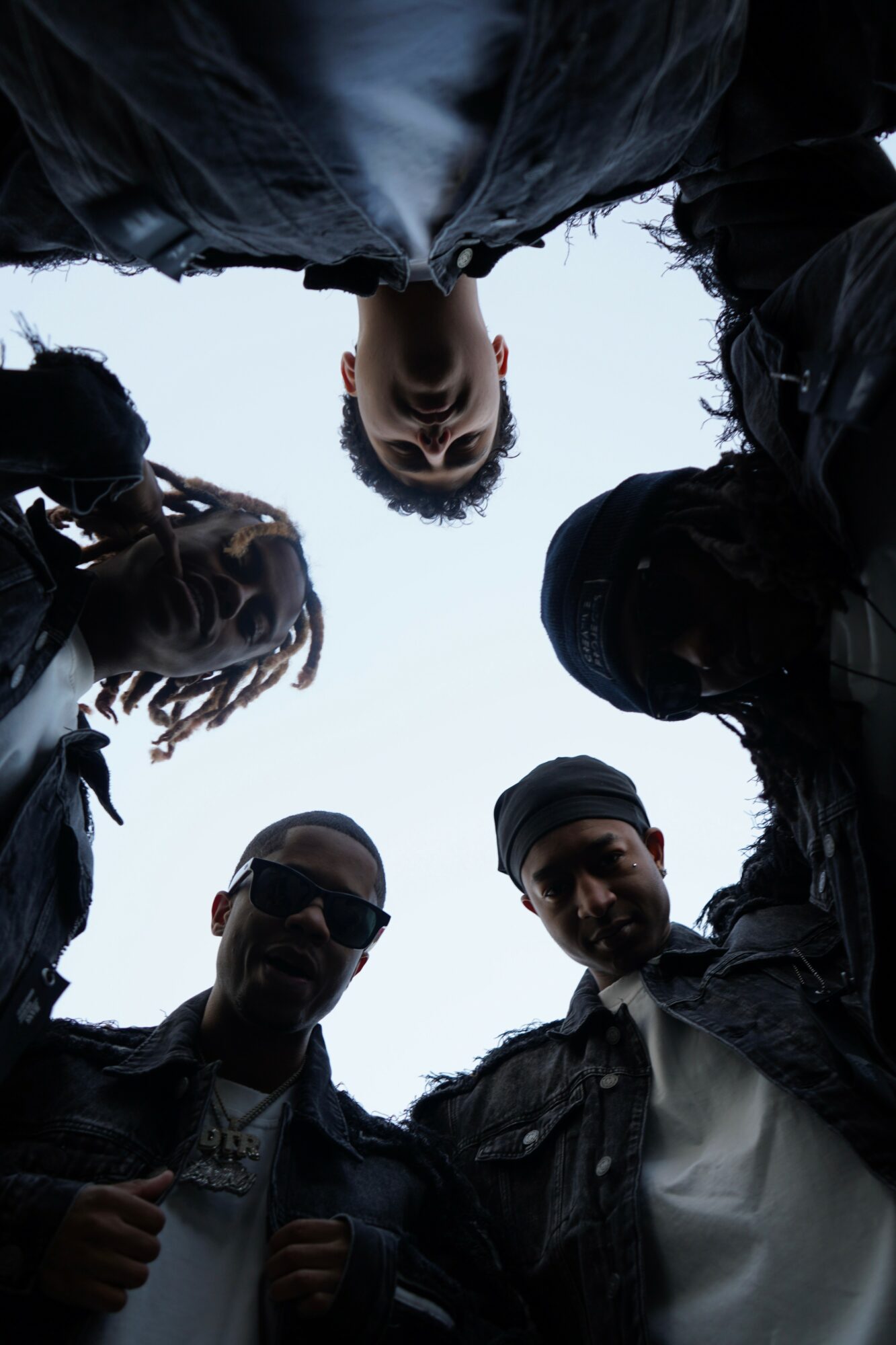 Group of five people looking down at camera, sky background, some wearing sunglasses and dark clothing.