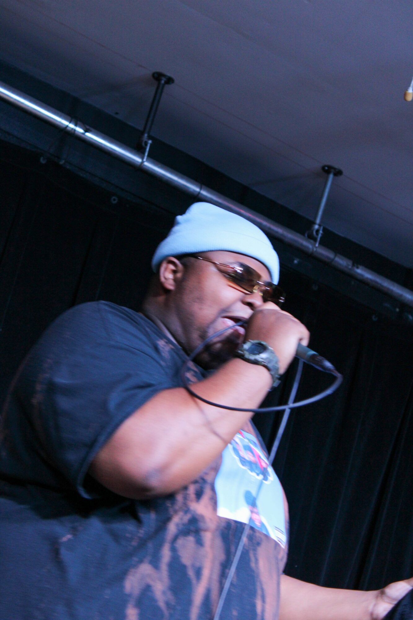 Person wearing a white beanie and sunglasses singing into a microphone on stage.