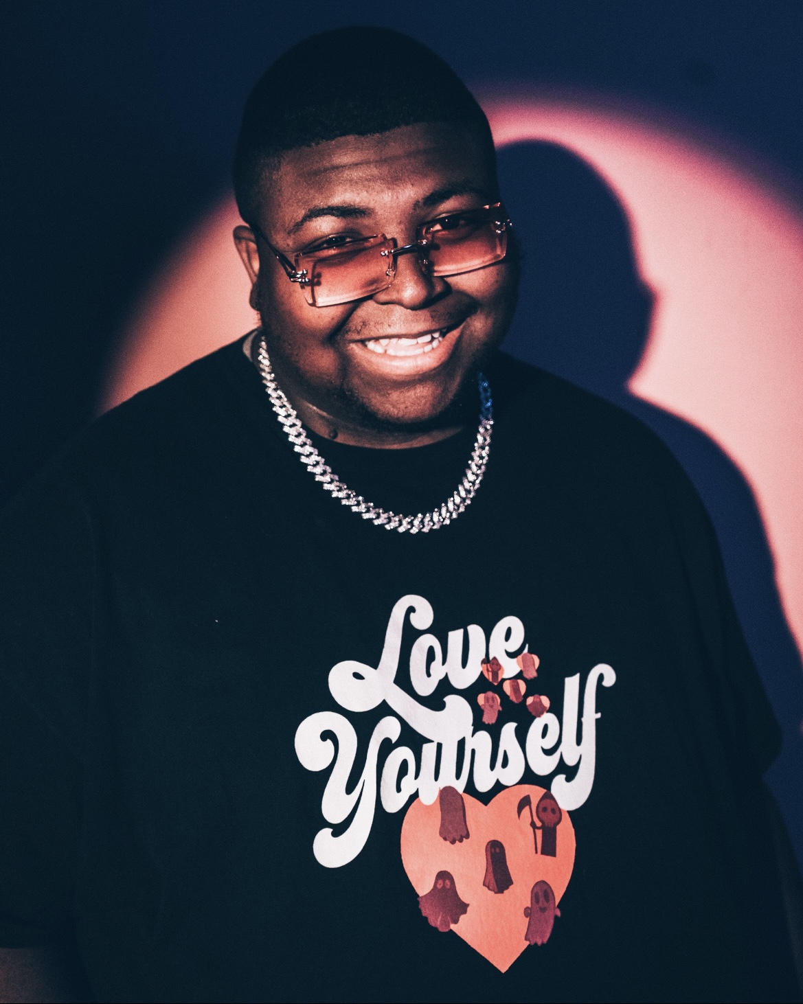 Young man smiling, wearing glasses, a chain necklace, and a black shirt with a heart and text, in a dark setting with pink light.