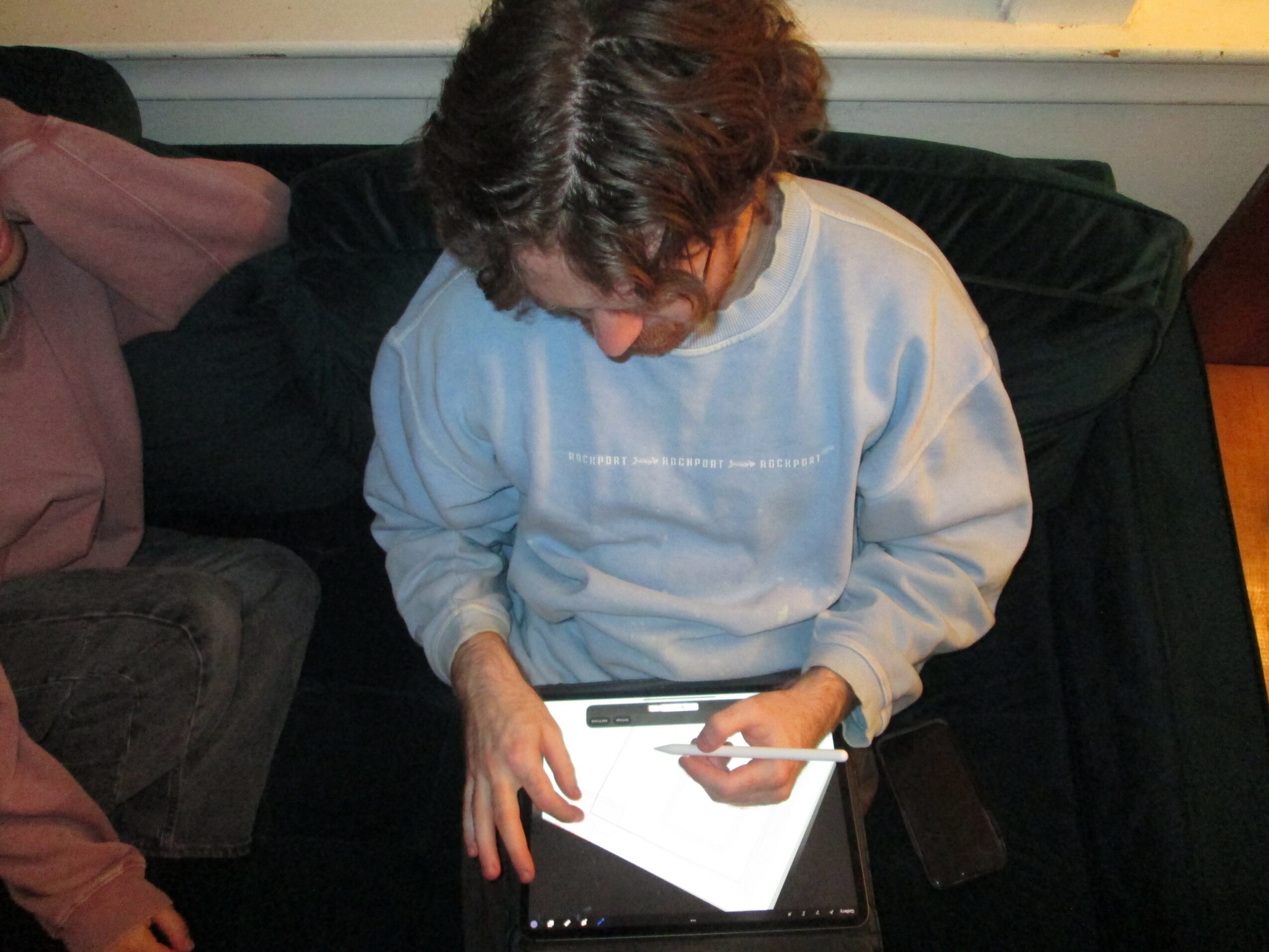 Person with short brown hair wearing a light blue sweatshirt, sitting on a black couch, writing on a tablet with a stylus.