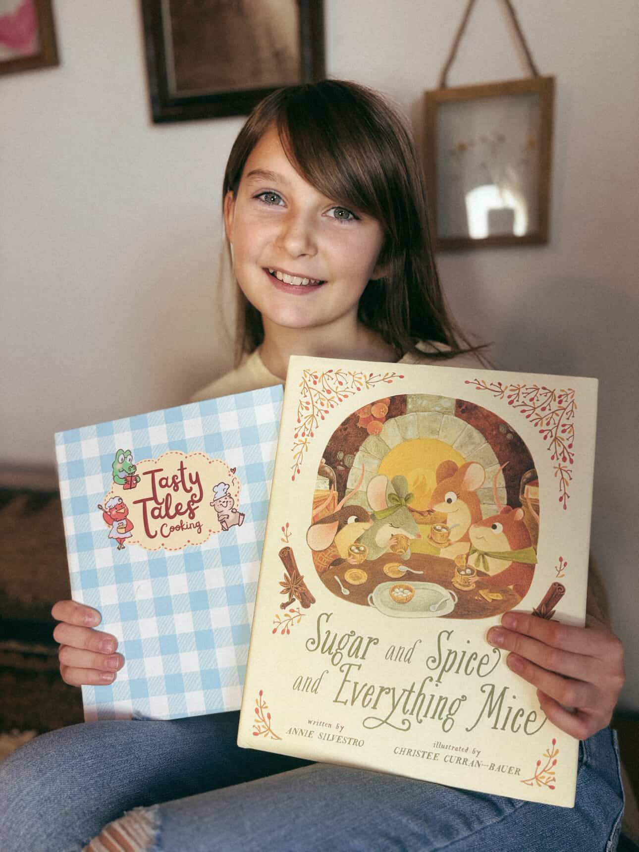 Young girl with brown hair holding two books, smiling, in a room with framed pictures on the wall.