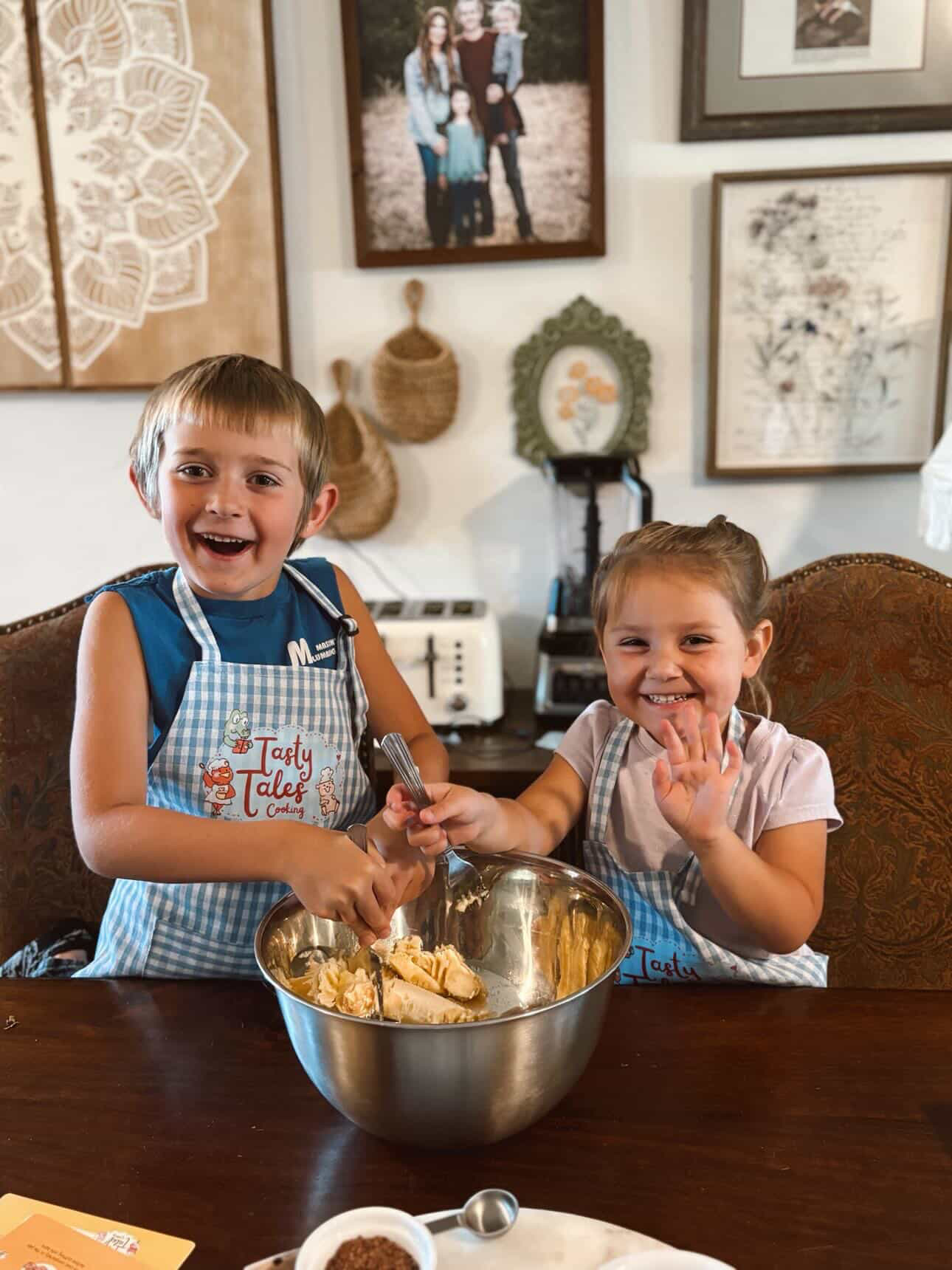 Two children, a boy and a girl, smiling and shaking hands over a mixing bowl in a kitchen, wearing aprons.