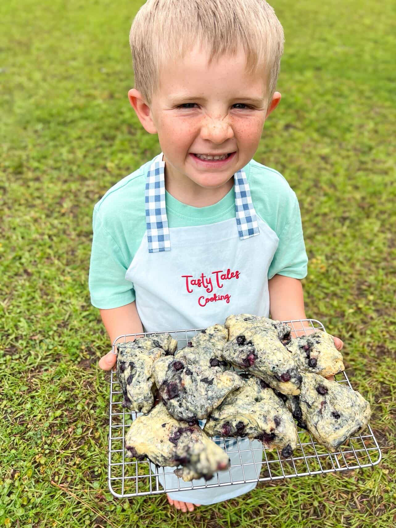 Young boy holding a tray of blueberry muffins outdoors on grass, smiling, wearing a light green shirt and apron.