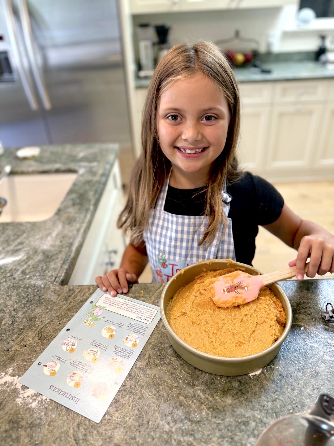 Young girl with long hair smiling in a kitchen, stirring a bowl of orange mixture, wearing an apron, with a recipe card nearby.