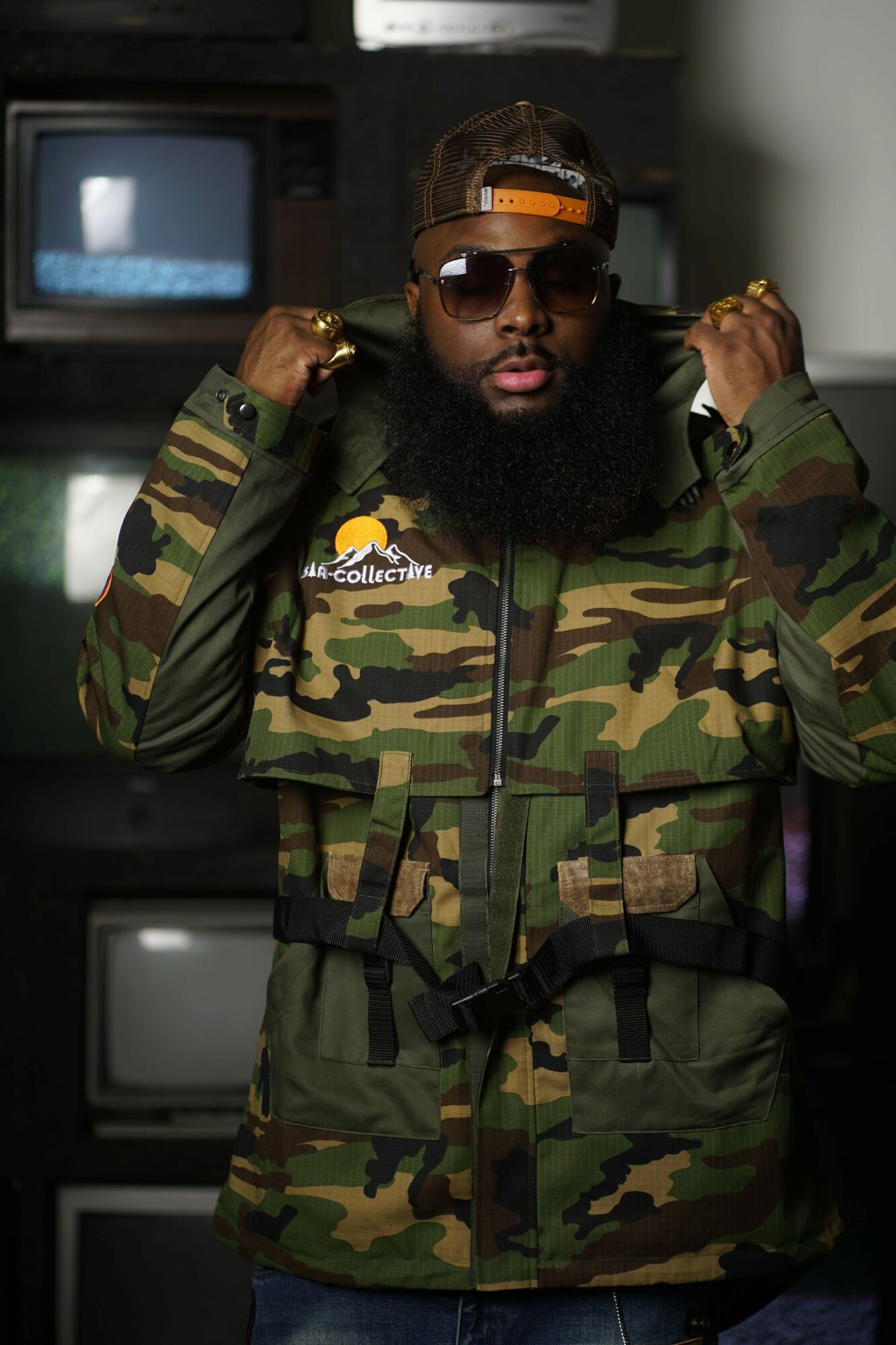 Man with sunglasses and a beard wearing a camouflage jacket, adjusting the hood, in a room with electronic equipment