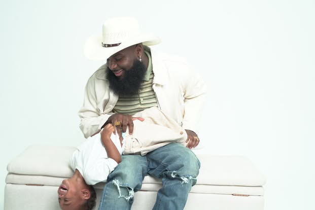 A man with a beard and a child are smiling and playing together on a white surface, with a plain white background.