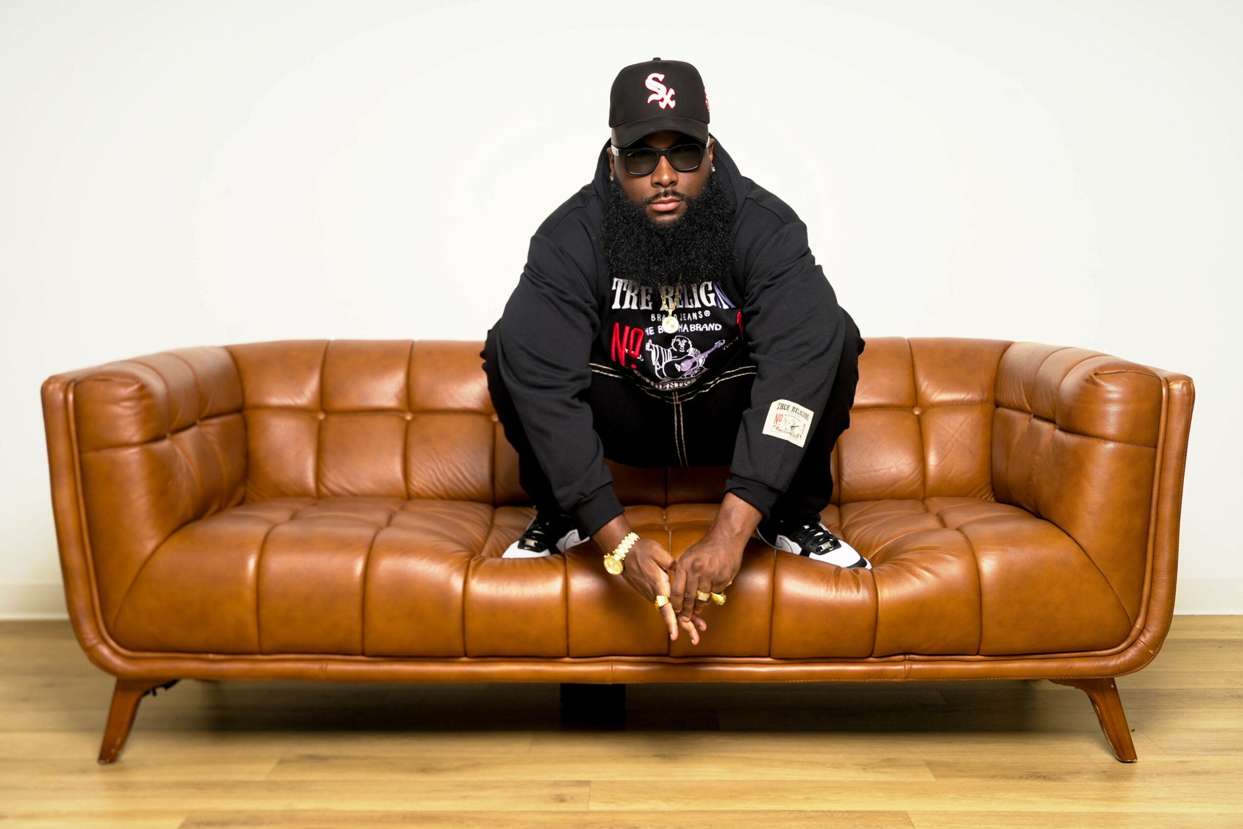 Person with a beard and sunglasses crouching on a brown leather sofa, wearing a black hoodie and cap, against a plain white background.