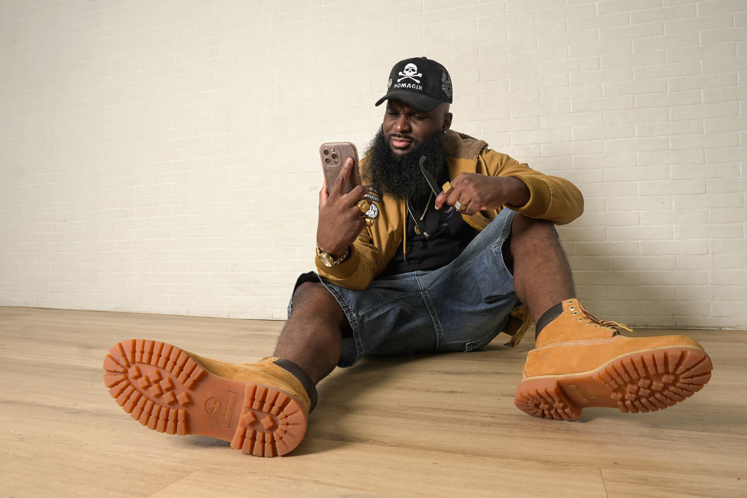 Person sitting on the floor taking a selfie, wearing a cap, jacket, shorts, and boots, against a plain wall.