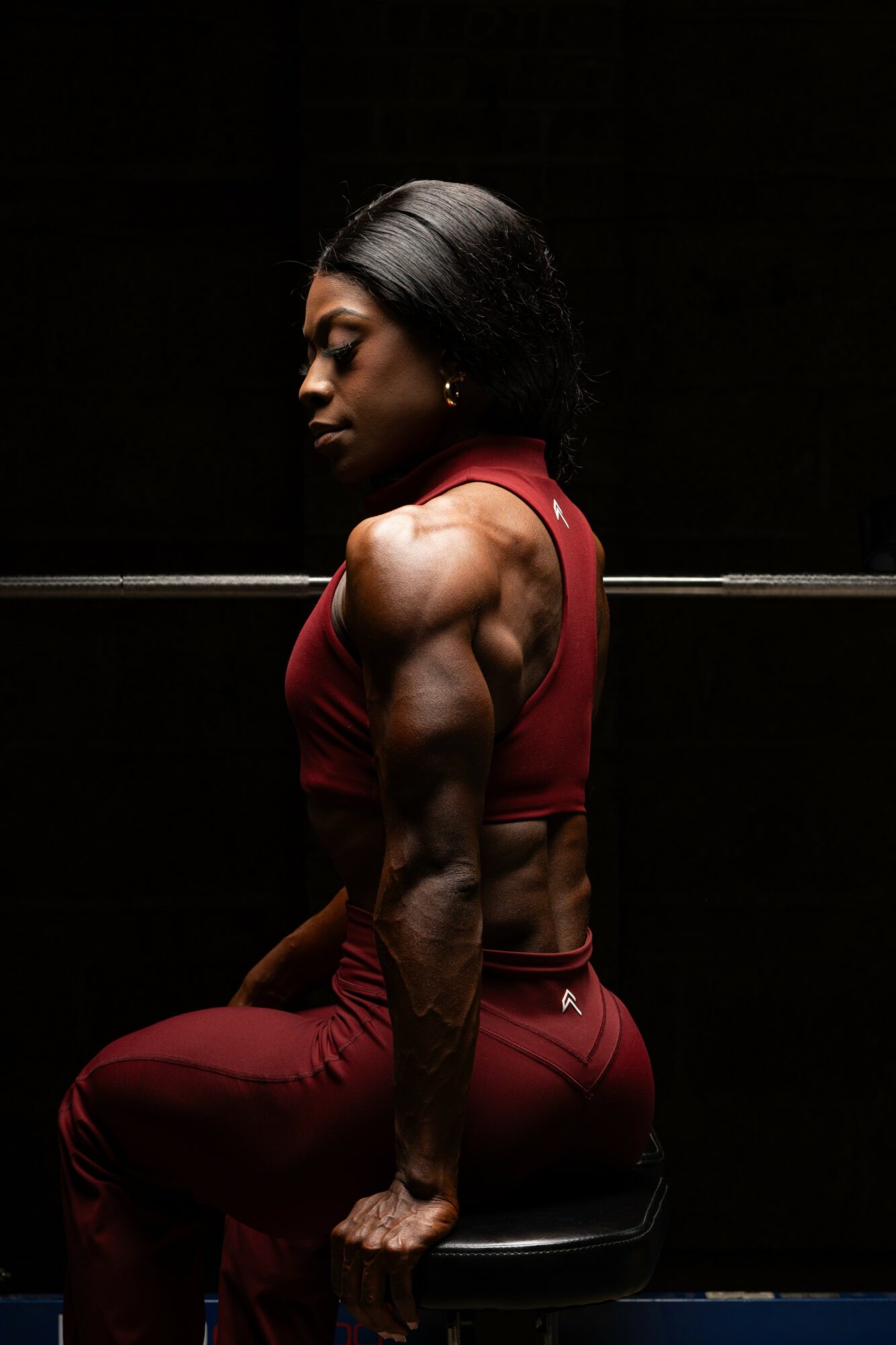 A muscular woman with dark hair in a red sports outfit, sitting on a stool in a dark setting.