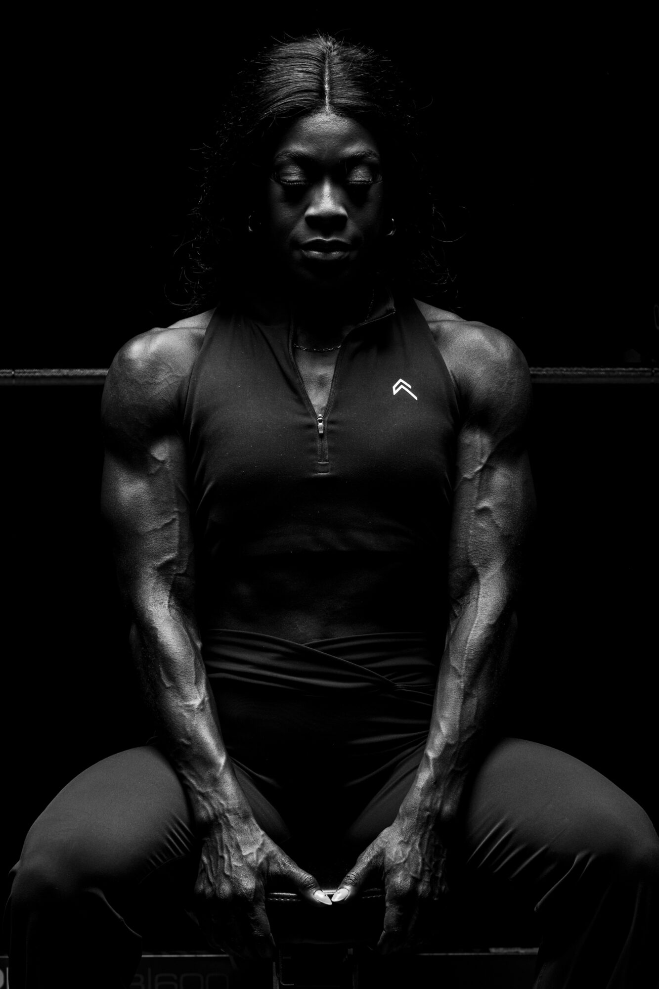 A muscular woman with long hair sits in a dark environment, looking serious, with arms resting on her thighs.