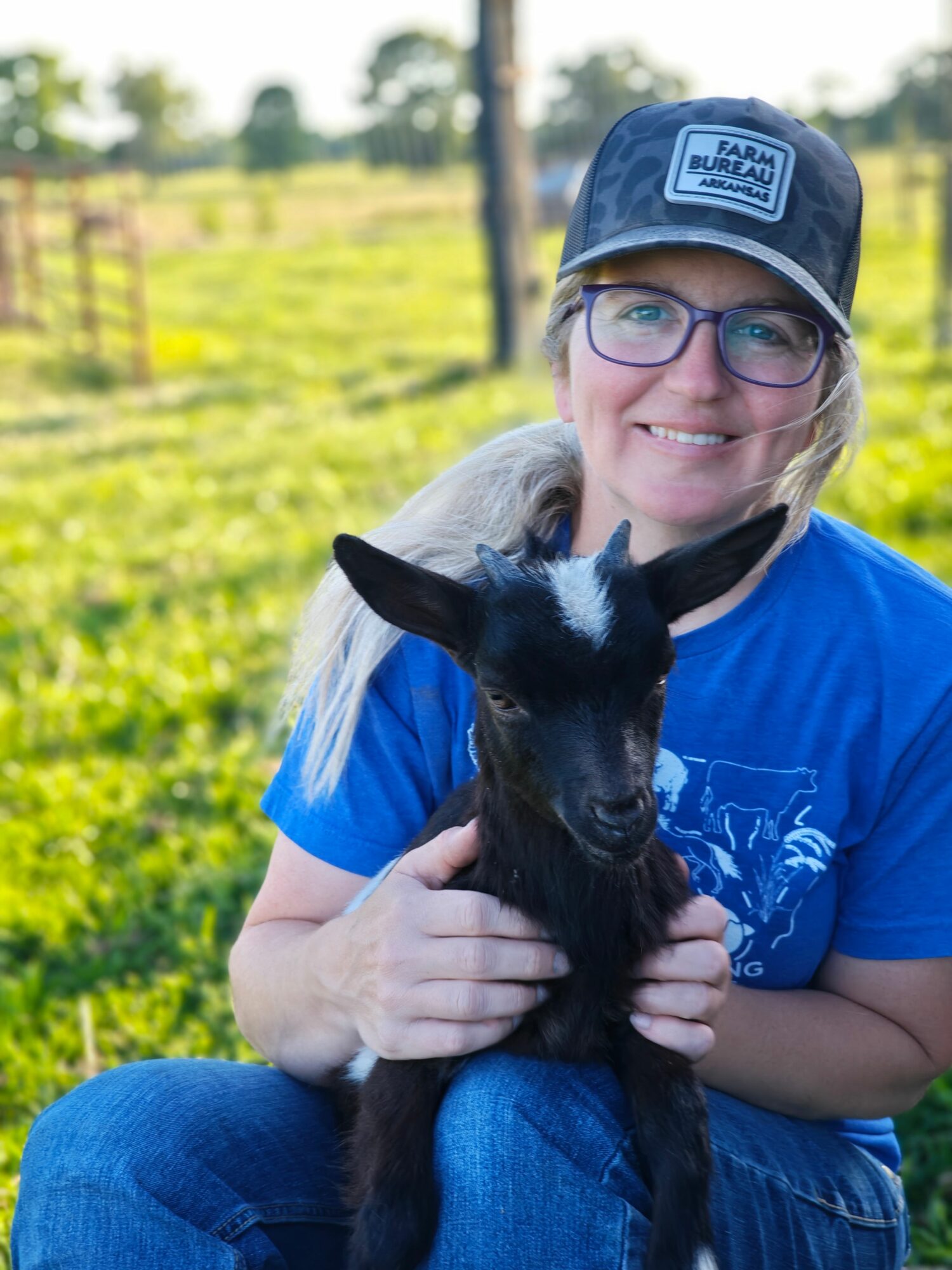 Person wearing glasses and a cap holding a black and white goat outdoors in a green field.