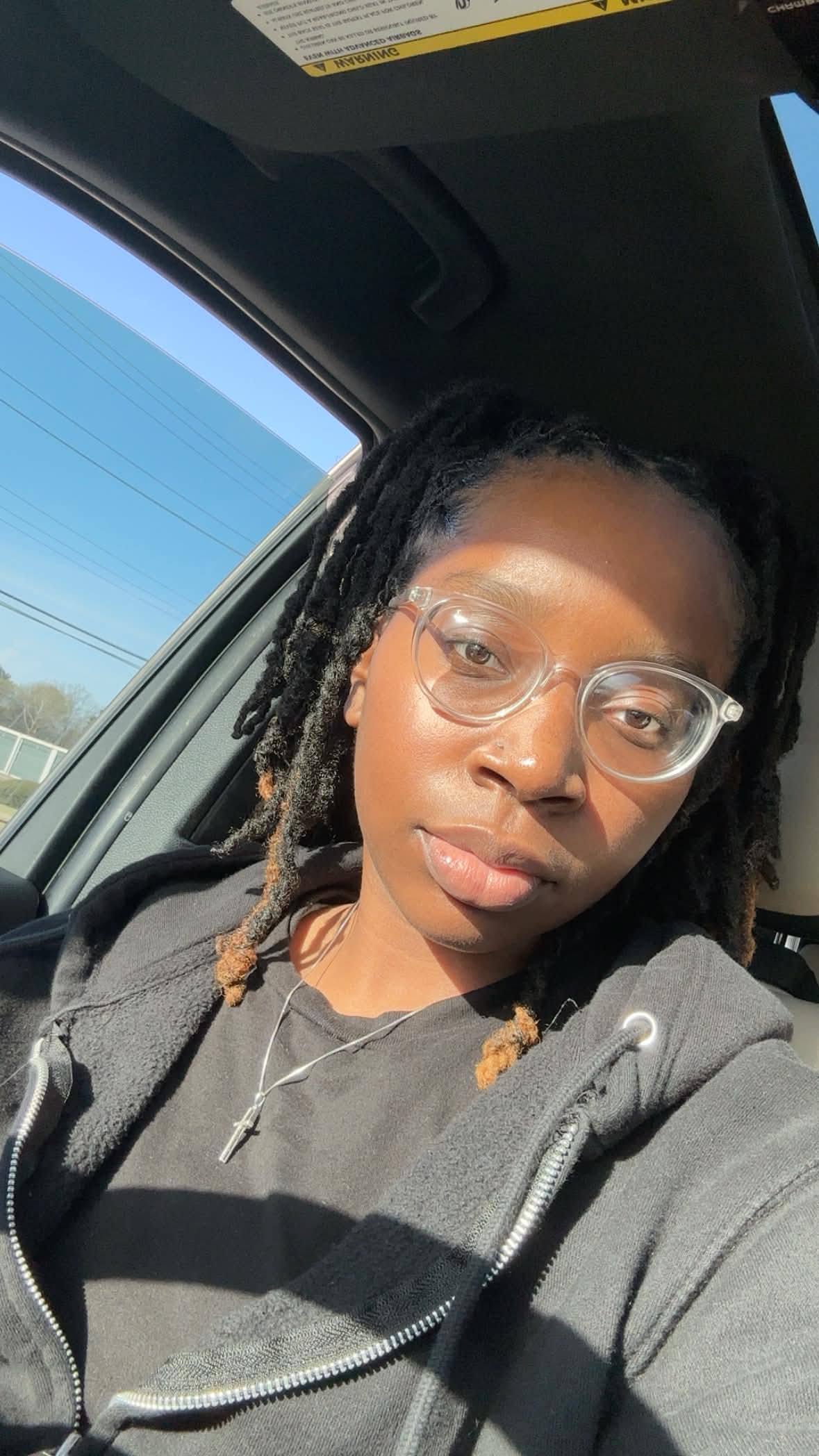 Young woman with glasses and braided hair sitting in a car, sunlight on her face, looking at the camera.