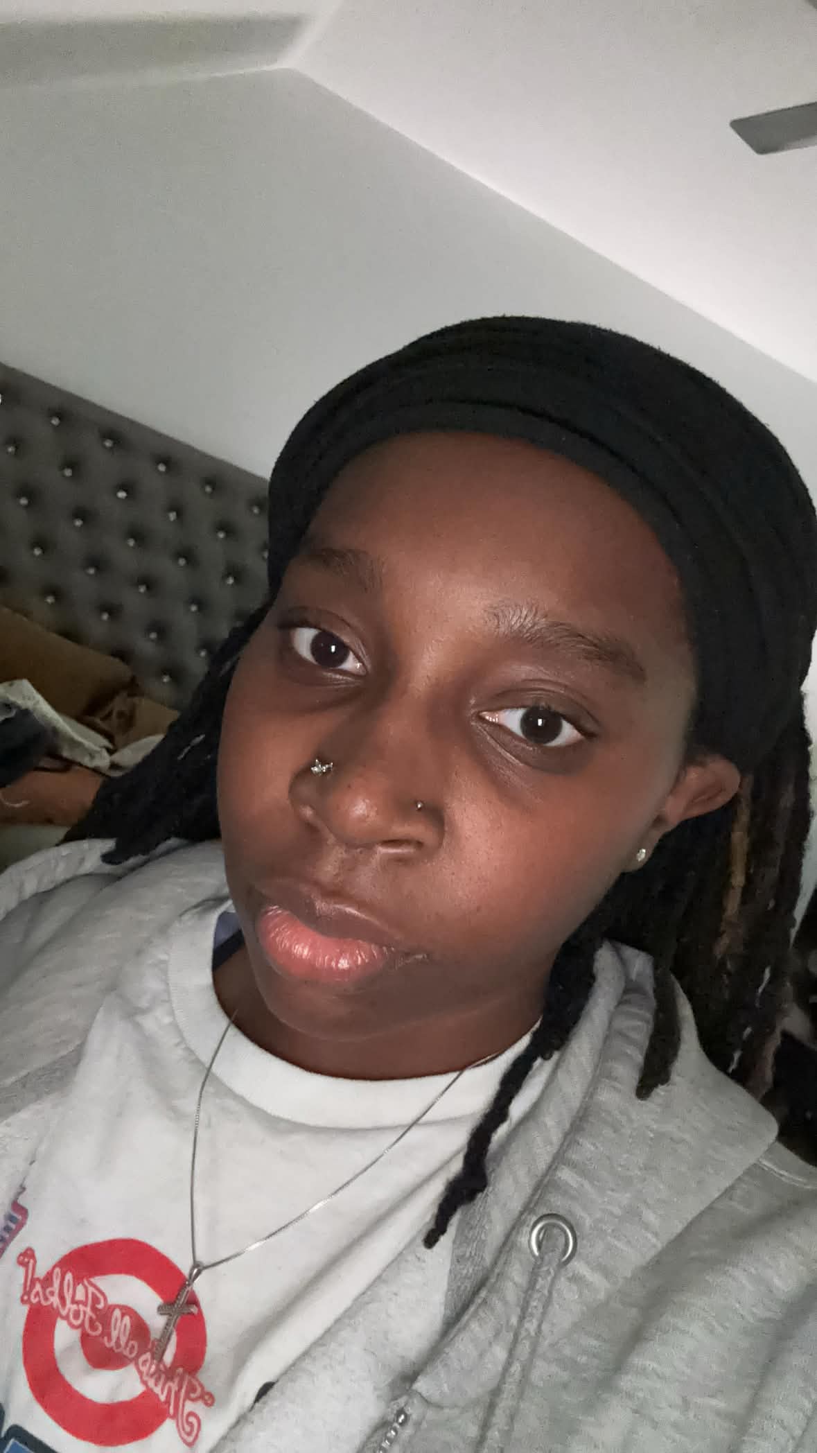 Young person with black headwrap and nose piercing, wearing a gray hoodie, in a room with a gray headboard.