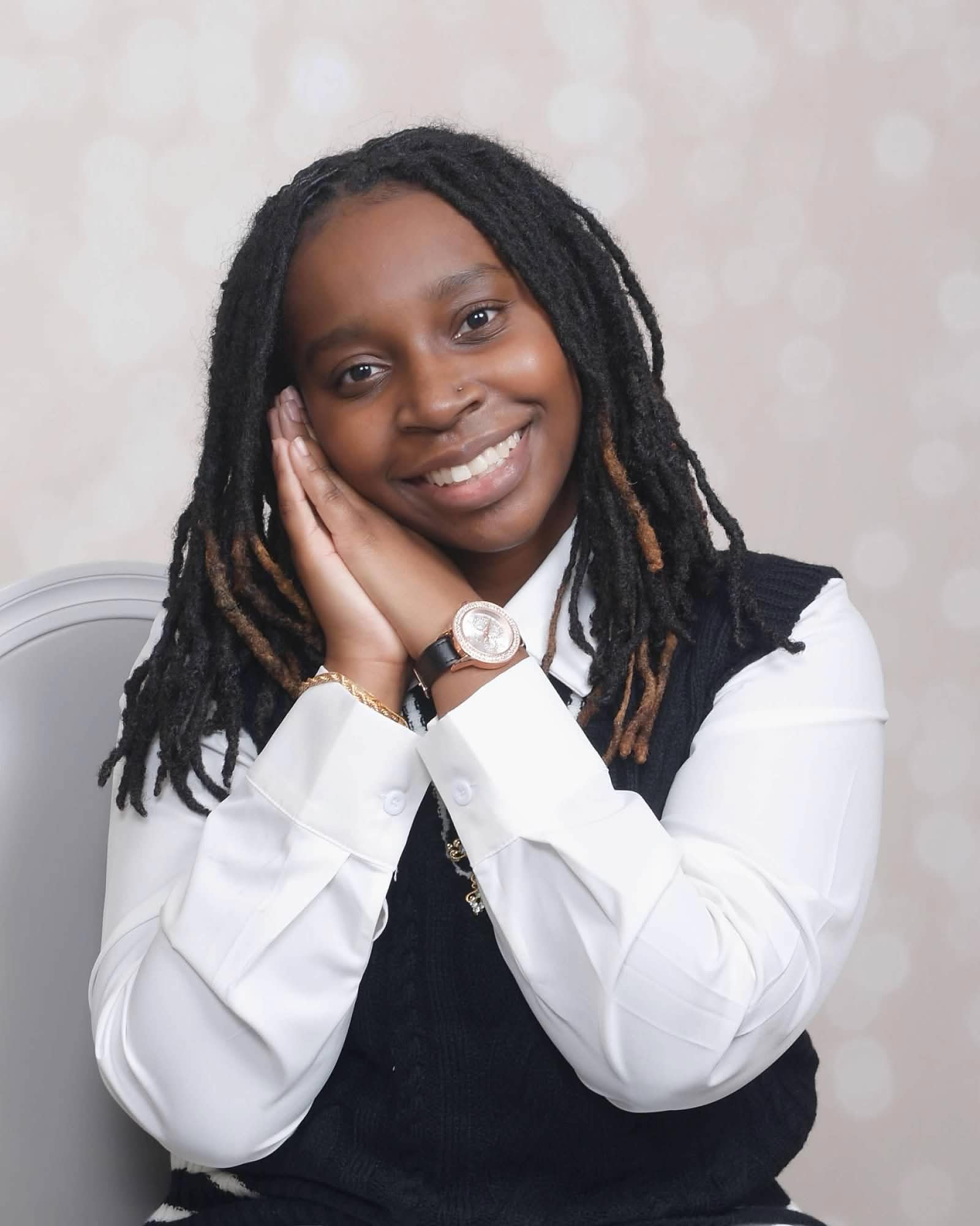 Young woman with dreadlocks smiling, resting her face on her hands, wearing a white shirt and dark vest, sitting against a light background.