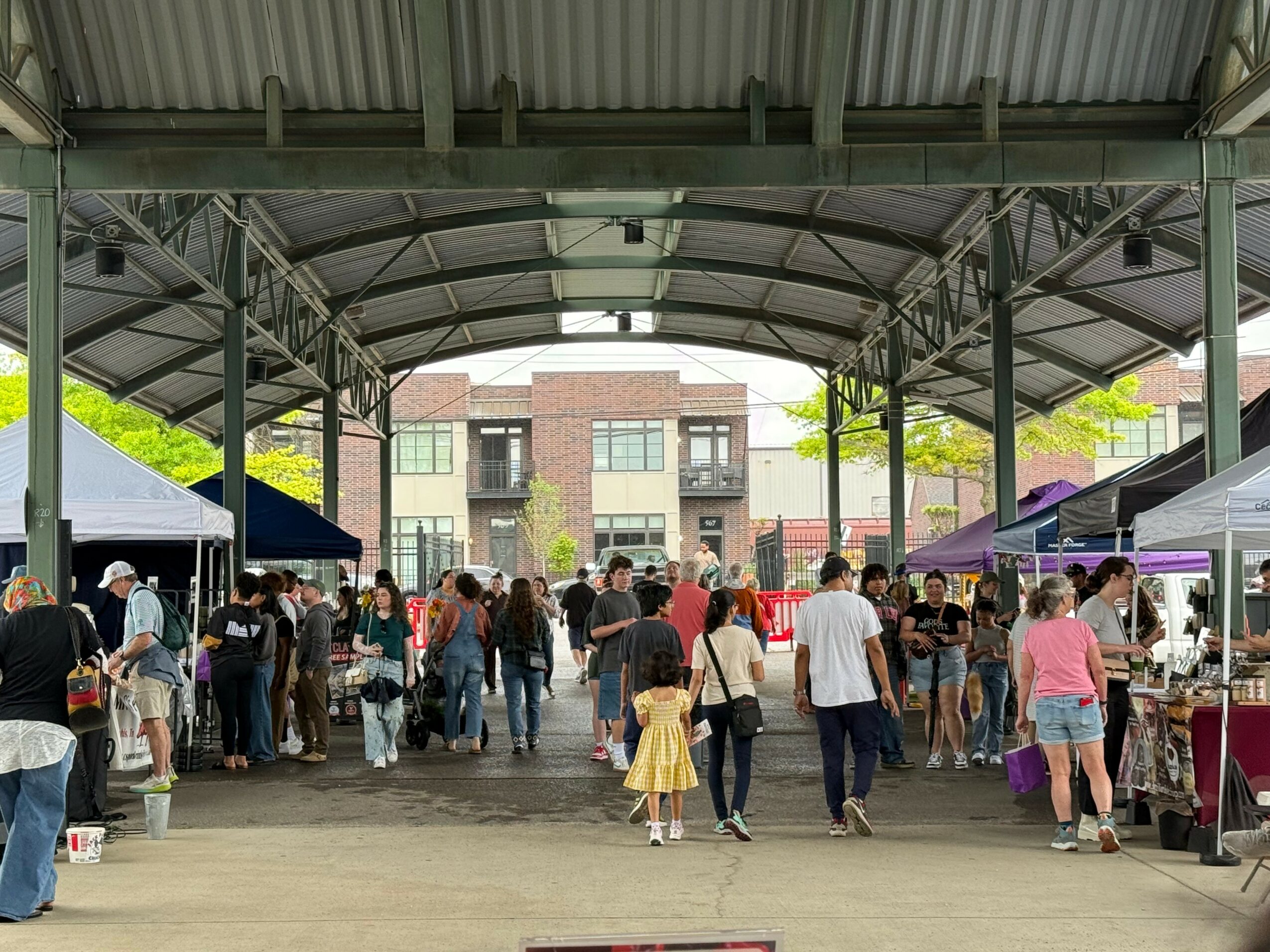 People walking through an outdoor market under a large covered structure with tents and stalls on both sides.