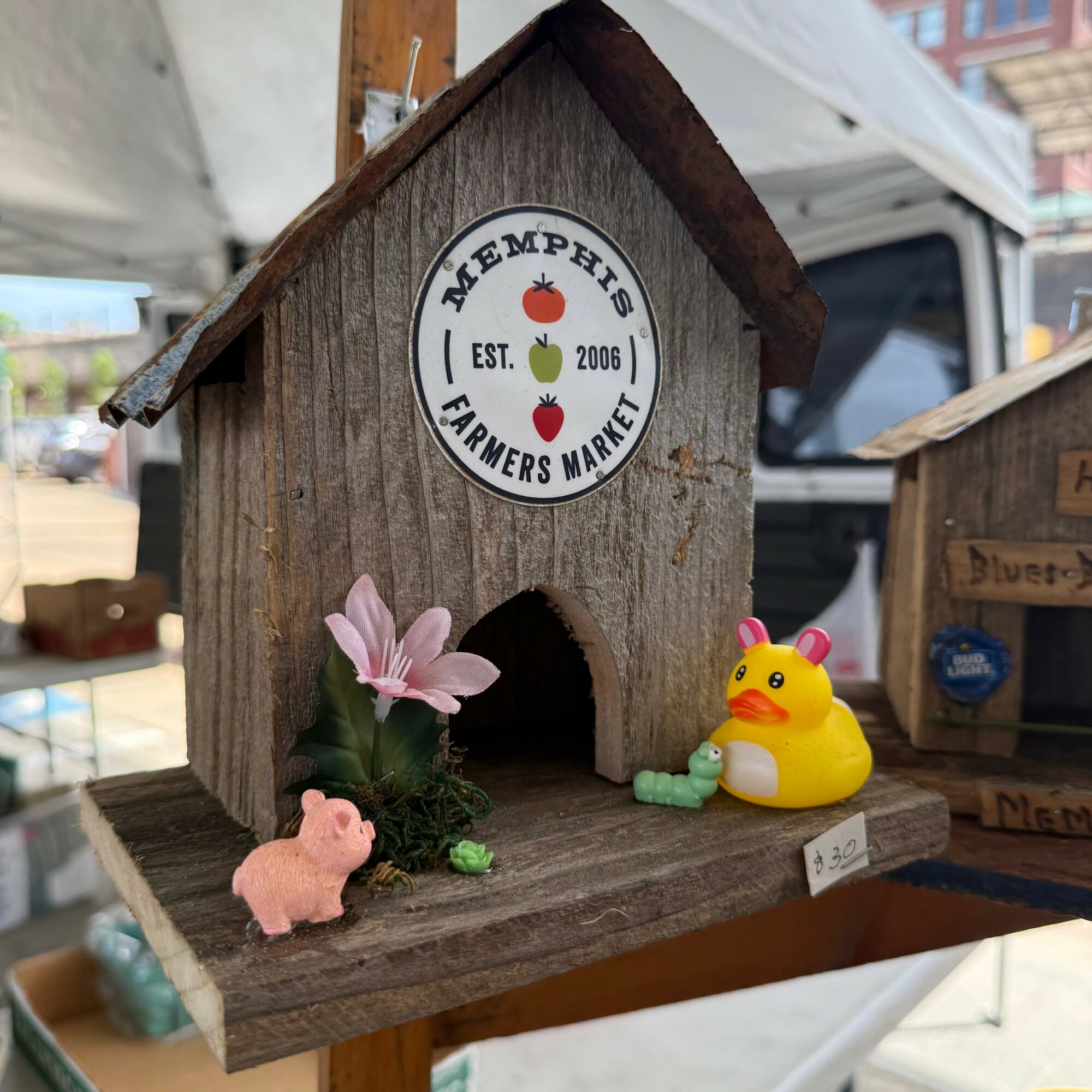 Wooden birdhouse with decorative items including a pink pig, a yellow duck, and a small plant, outdoors at a market stall.