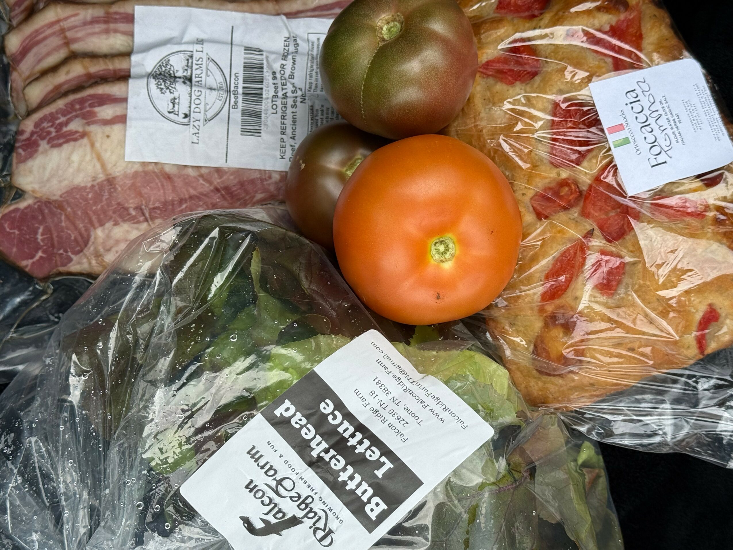 Tomato, two green tomatoes, lettuce in plastic bag, bacon, and bread in plastic packaging on surface.