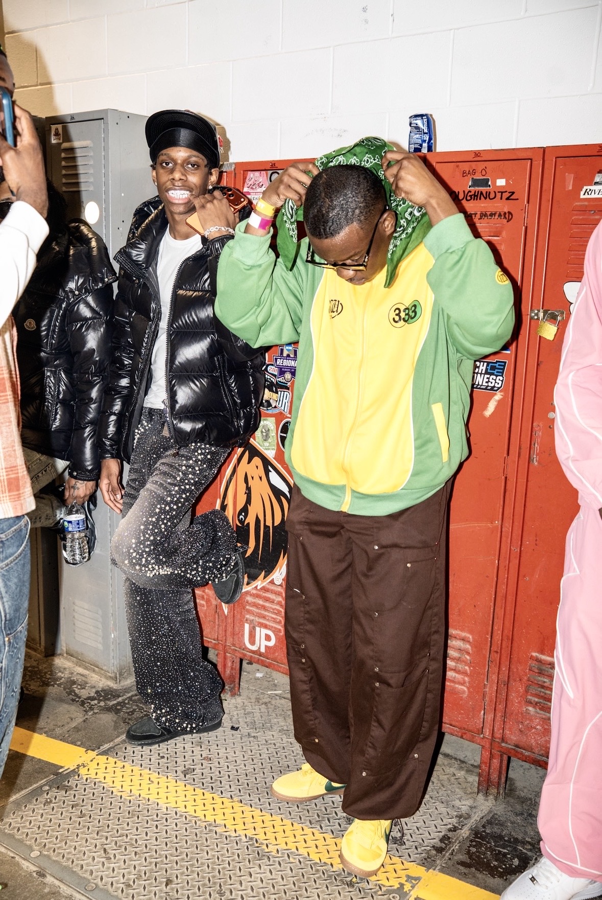 Two people standing in front of red lockers, one adjusting their hoodie, the other smiling and looking at the camera.