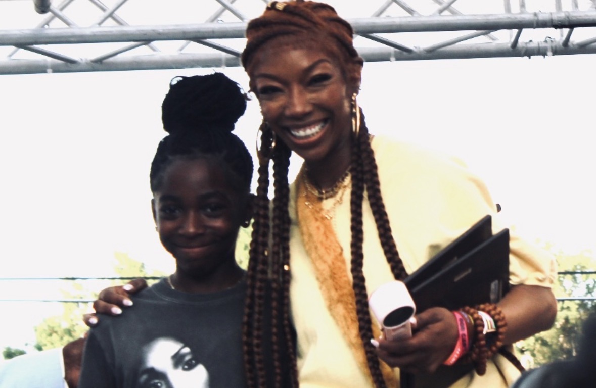 Two women smiling, one with braided hair holding a microphone and folder, the other with a bun hairstyle, outdoors.