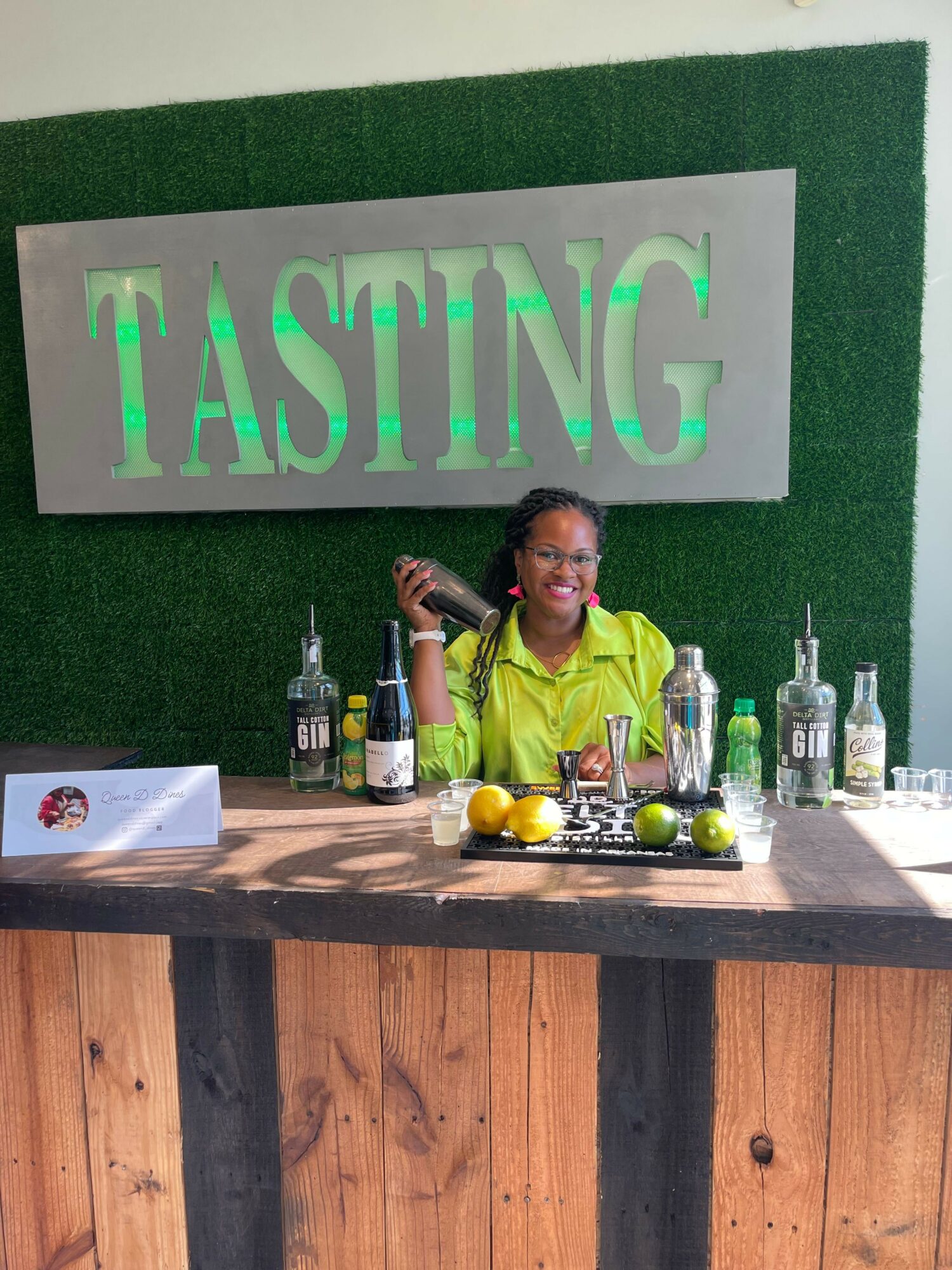Person behind a bar with various bottles and fruits, standing in front of a green wall with a sign reading 'TASHING'.