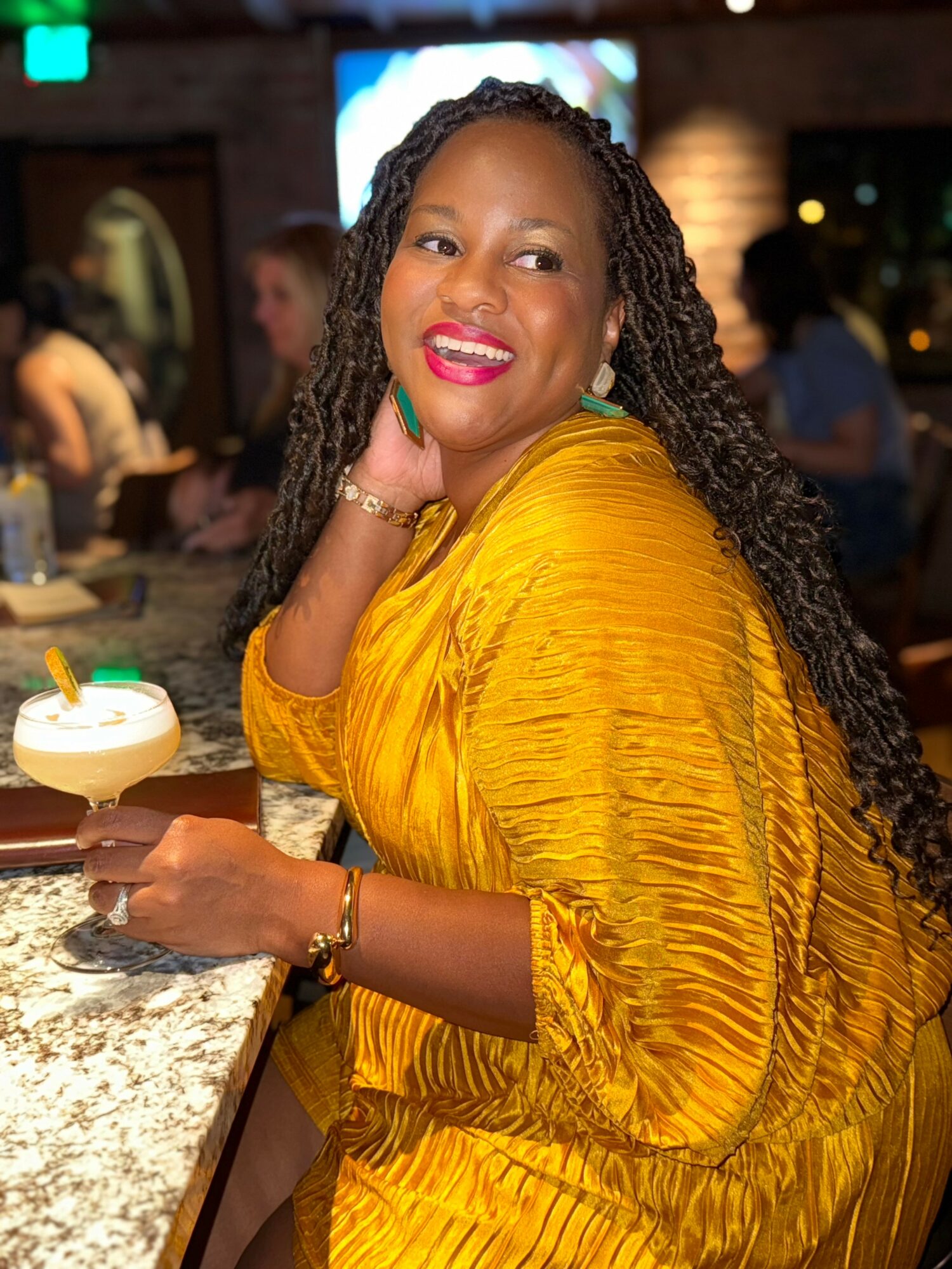 Smiling woman with long curly hair in a yellow dress sitting at a bar counter, holding a cocktail, in a lively indoor setting.
